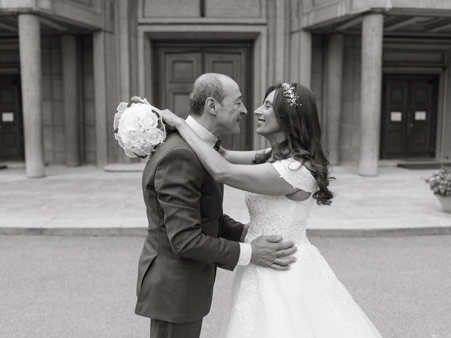 Black and white wedding photo of bride and groom embracing outdoors near elegant building columns.