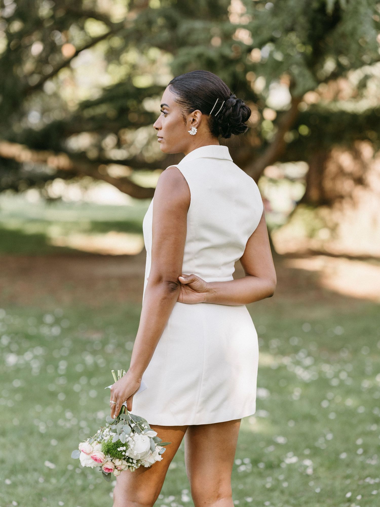 Bride in chic white sleeveless blazer mini dress viewed from behind, holding bouquet in sunny park.
