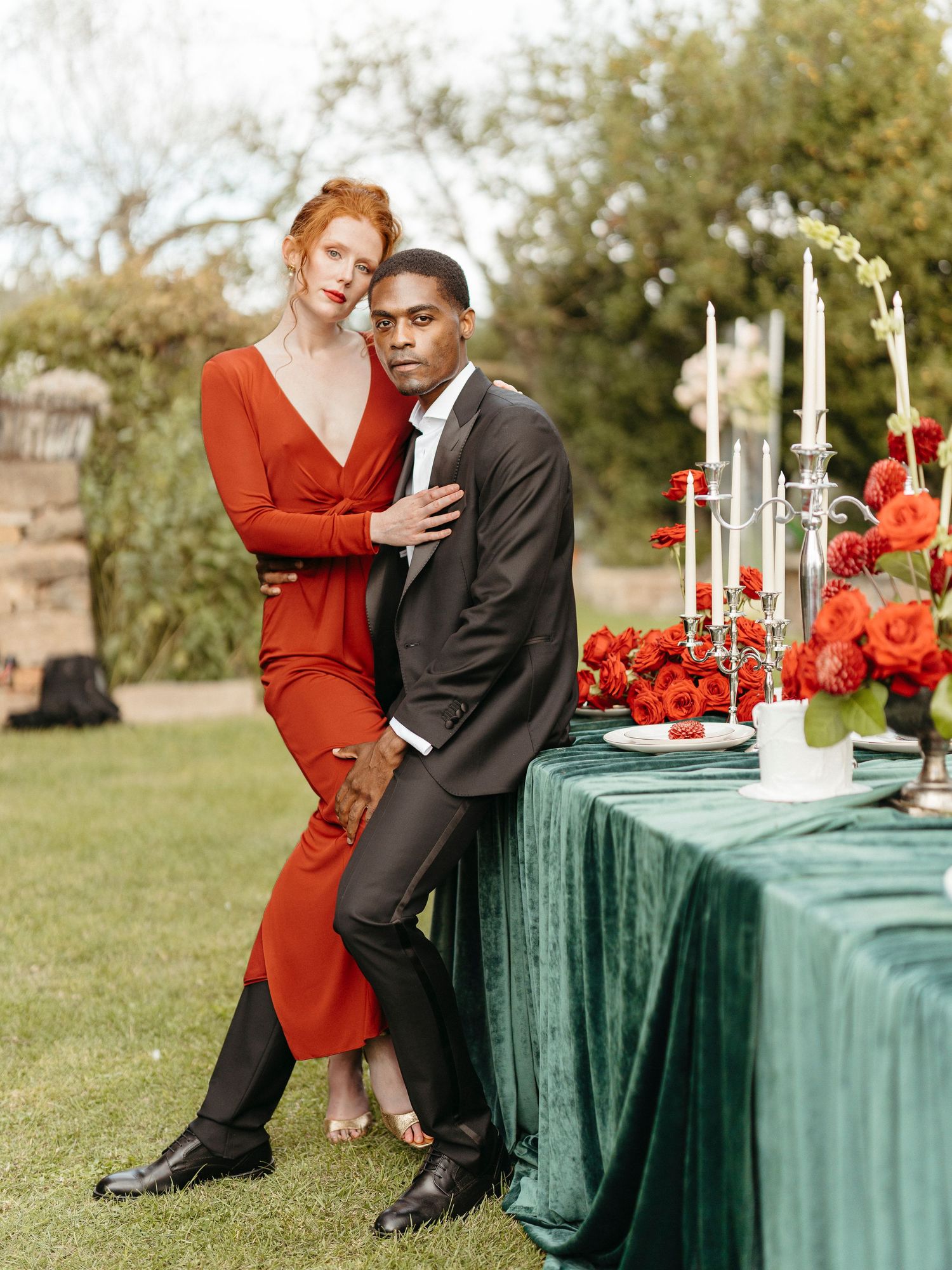 Couple poses elegantly beside a green velvet-draped table adorned with red roses and candelabras outdoors.