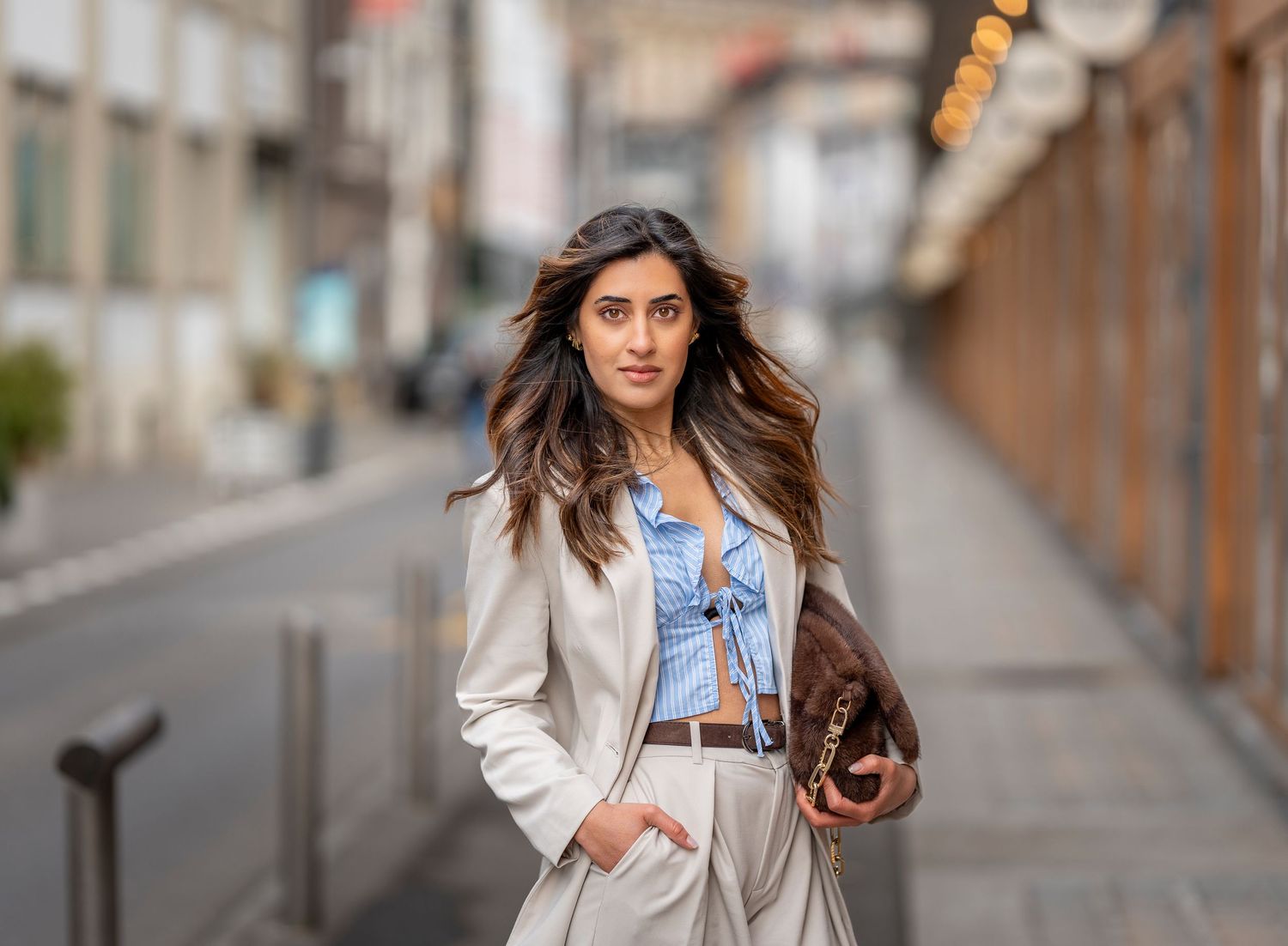 Confident woman in beige blazer and blue top walks along city street carrying brown handbag.