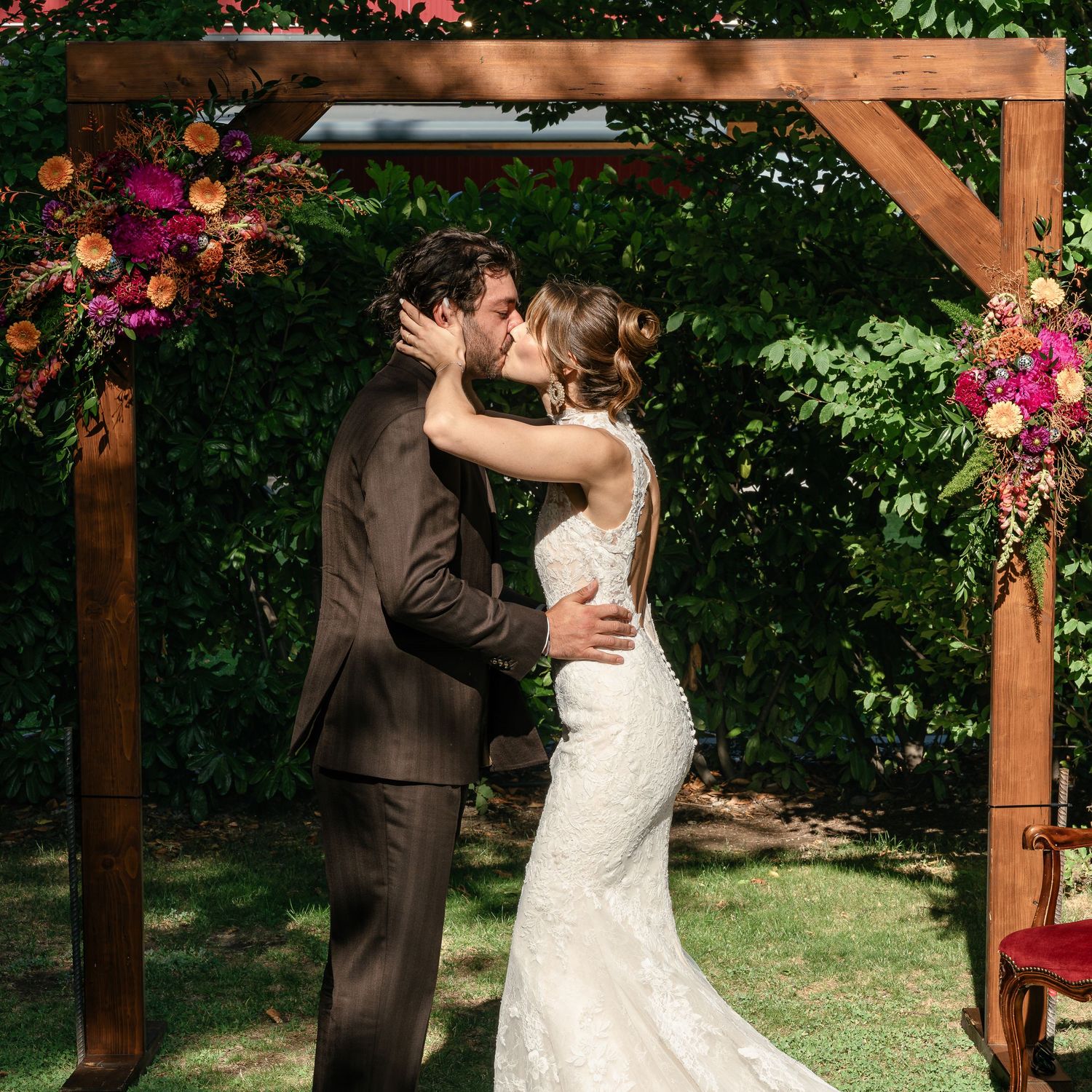 Bride and groom share first kiss under wooden floral arch with pink and orange blooms at outdoor garden wedding ceremony.