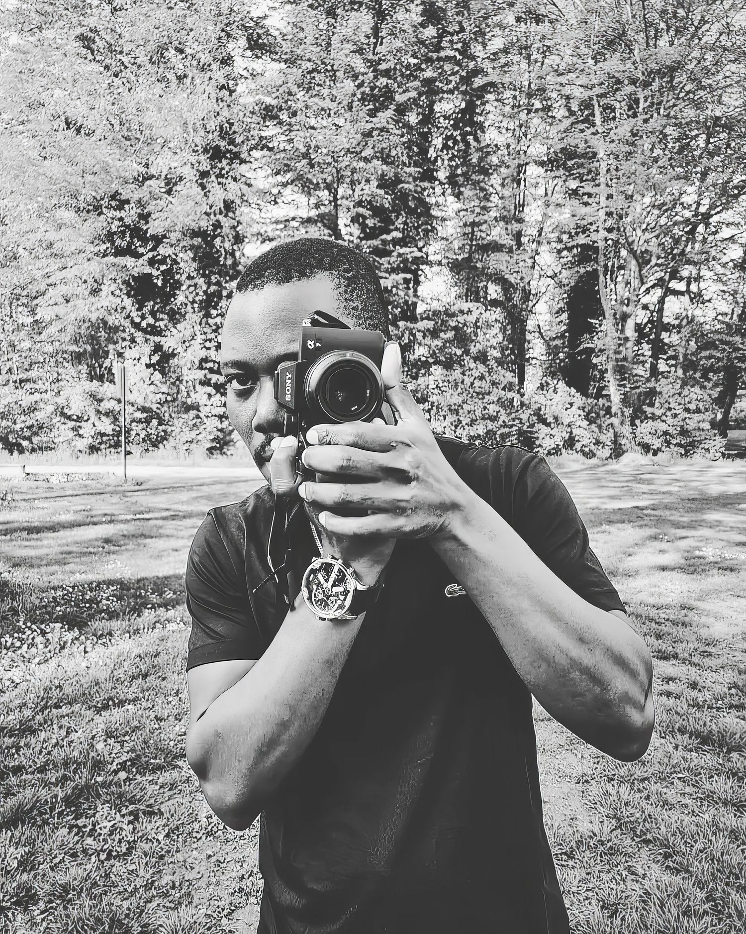 Man holding camera up to face photographing in black and white portrait among lush green trees.