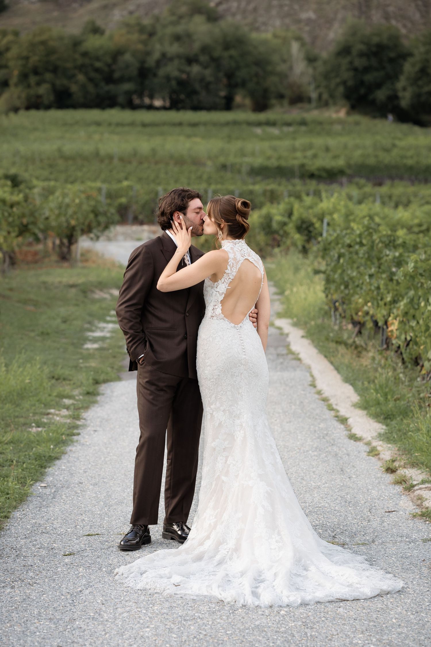 Bride and groom share a romantic kiss on a vineyard path, bride in lace backless gown, groom in brown suit.