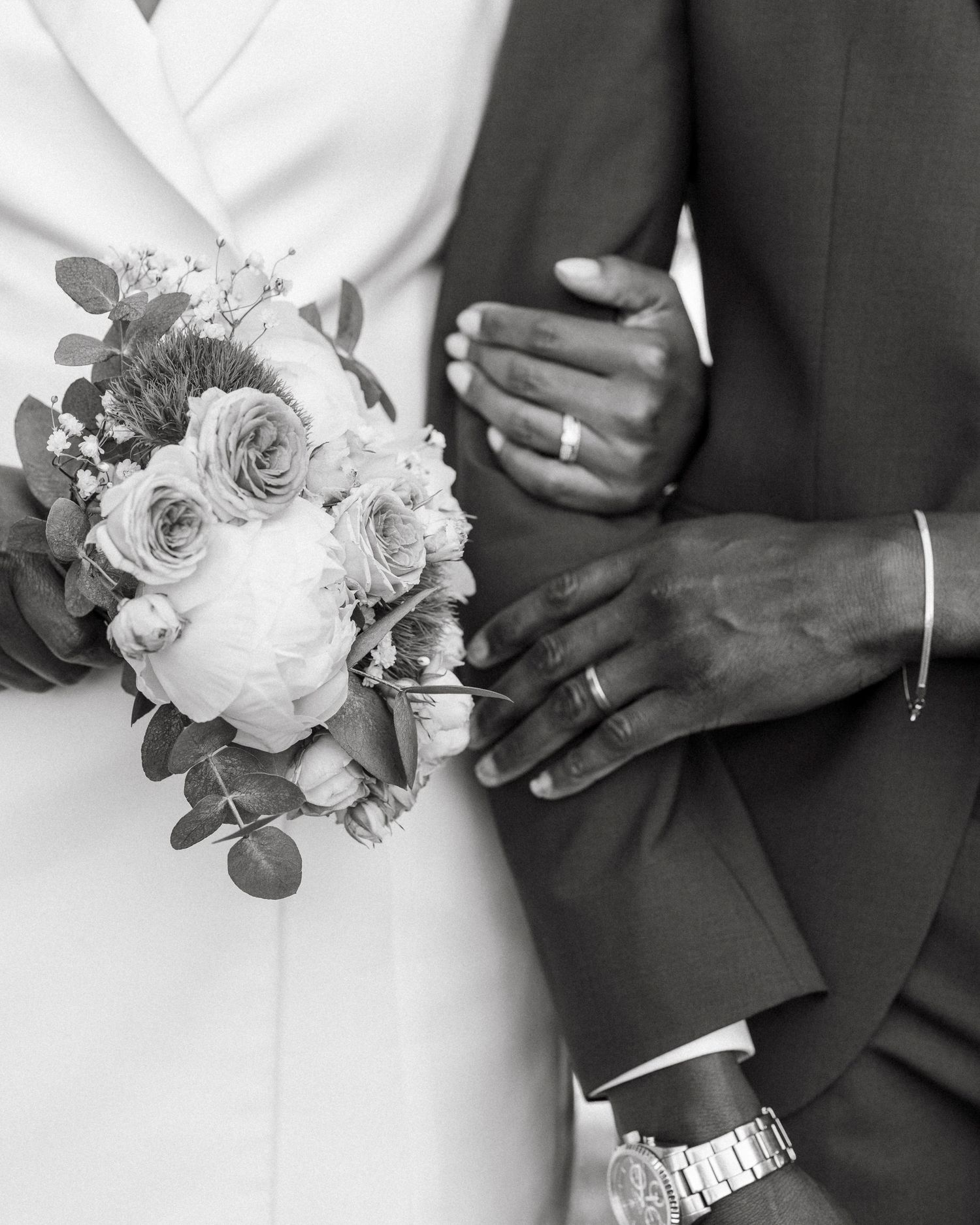 Black and white close-up of newlyweds hands showing wedding rings, bride holding floral bouquet.