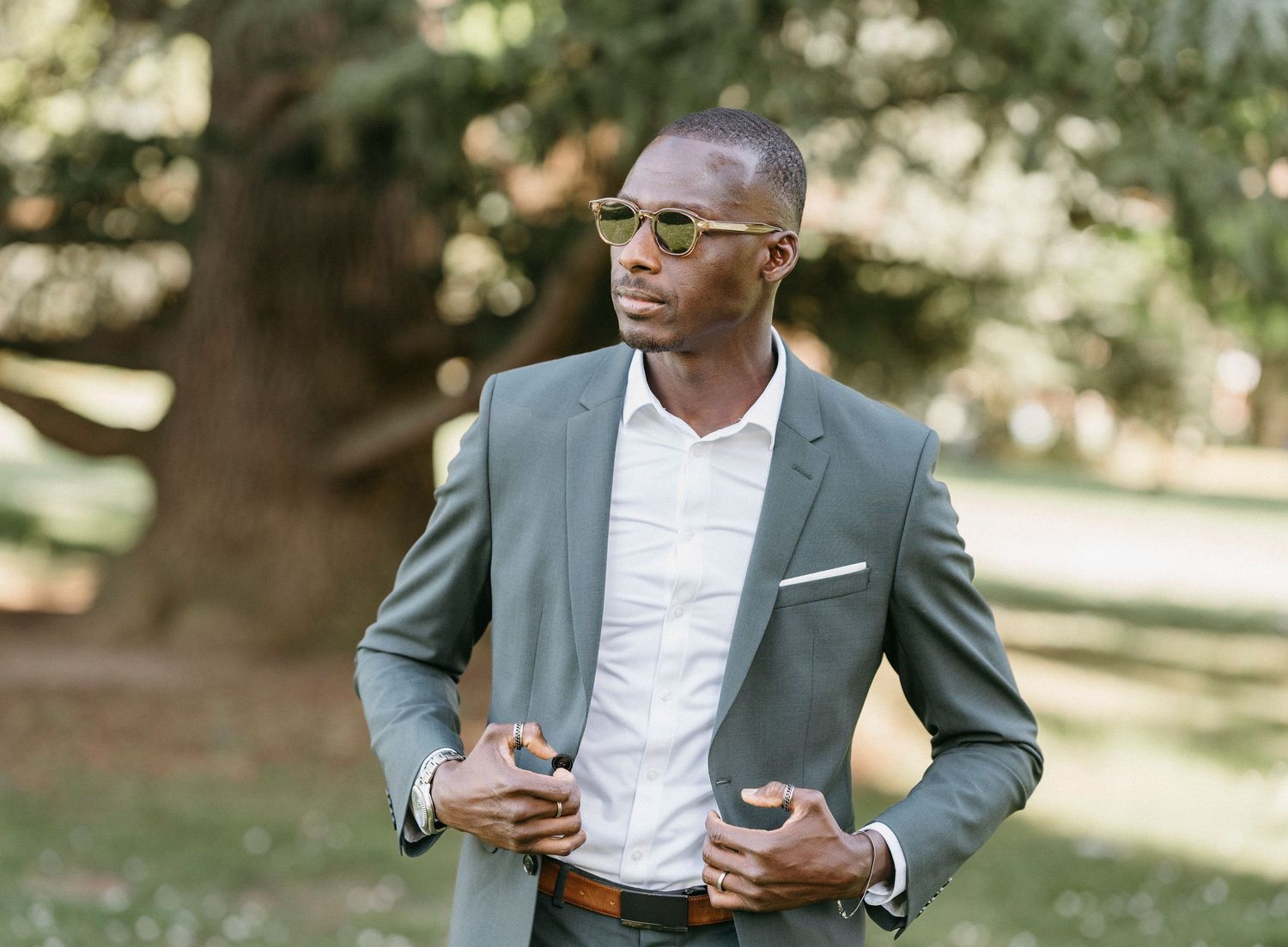 Confident groom in grey suit wearing green-tinted sunglasses, adjusting jacket in sun-dappled park setting.
