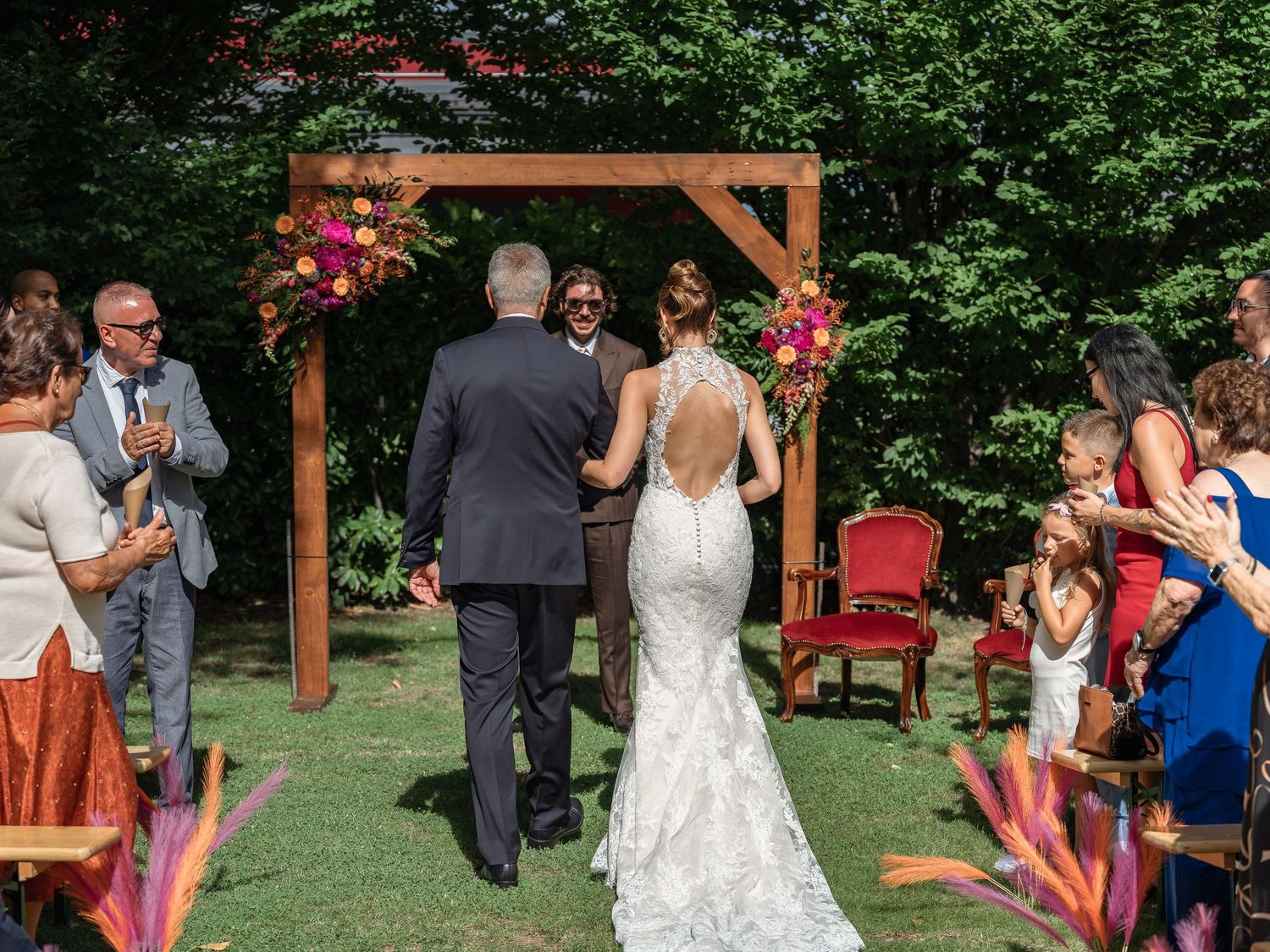 Bride walks down outdoor aisle with groom under wooden arch adorned with colorful flowers at garden wedding ceremony.