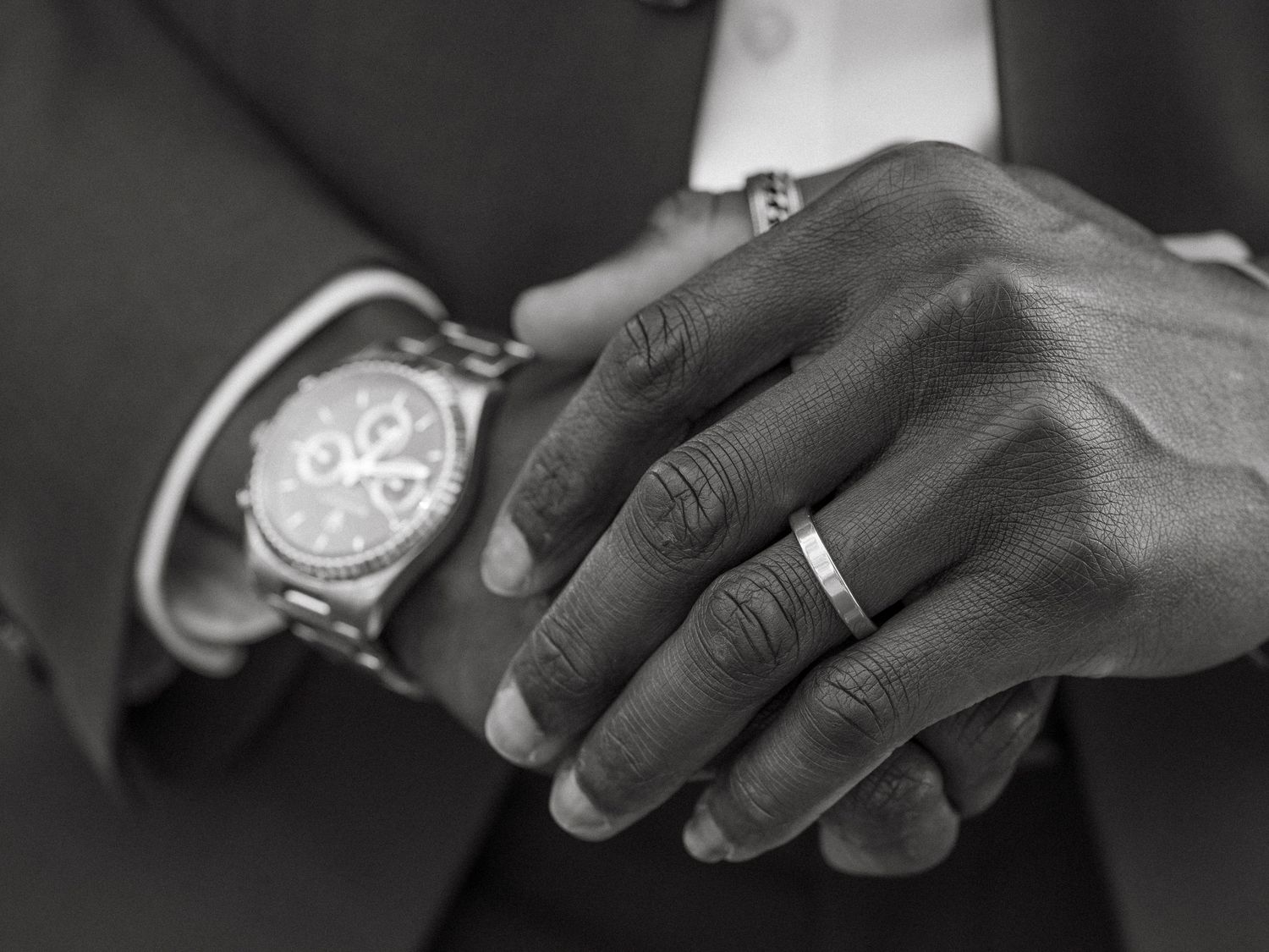 Close-up of a man's hands showing a luxury chronograph watch and a wedding band ring in black and white.