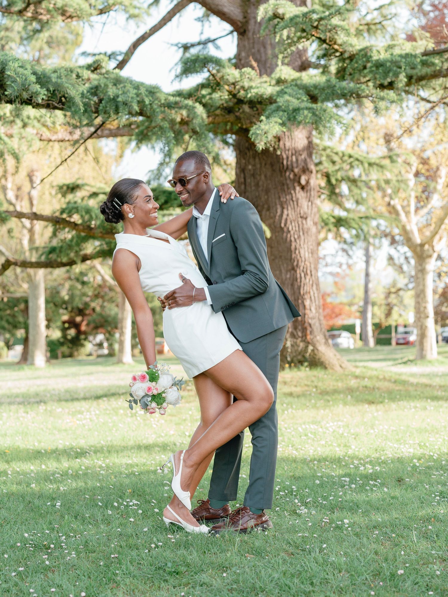 Smiling newlywed couple posing together under a large tree in a park, bride in white mini dress and groom in gray suit.