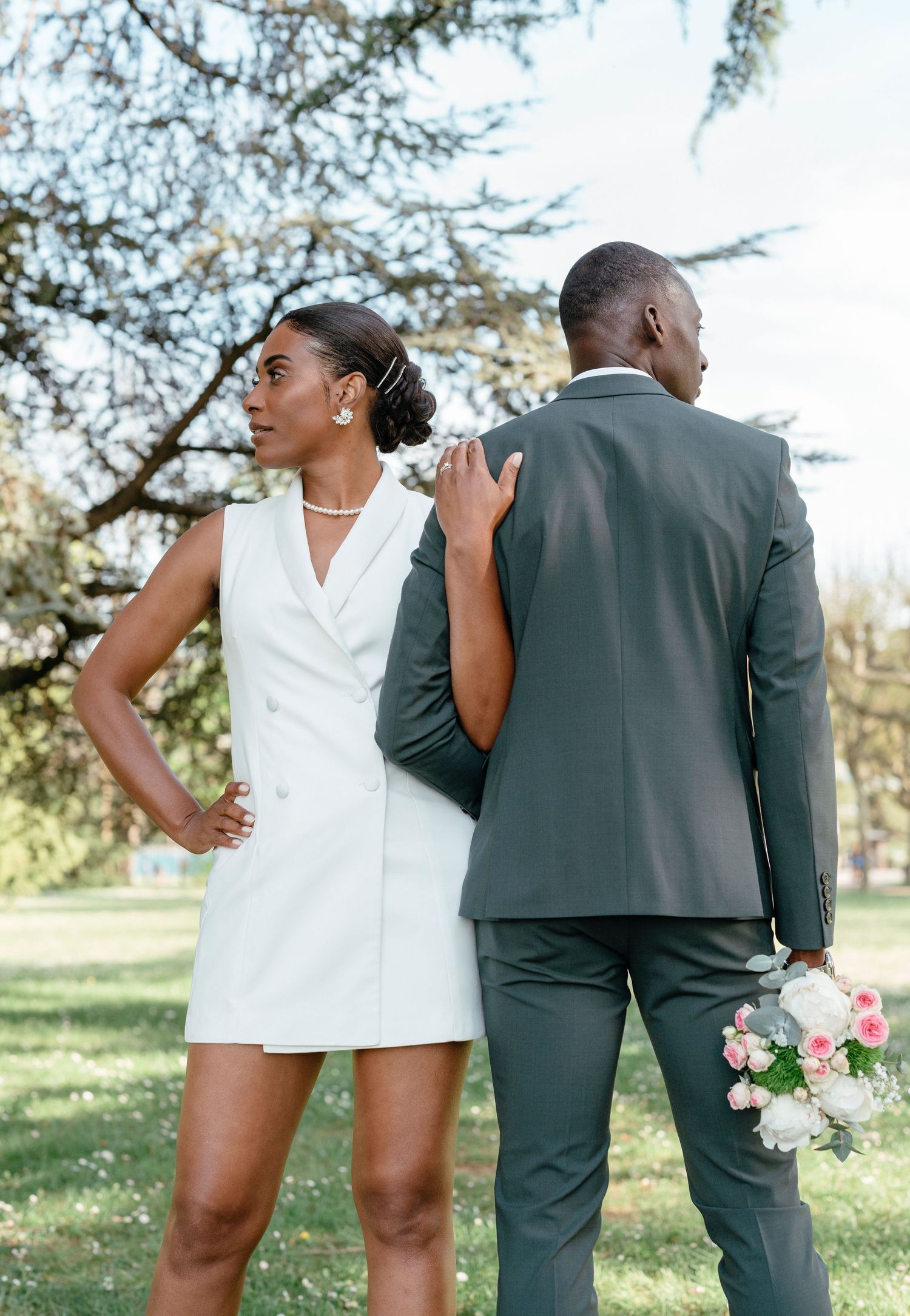 Bride in white tuxedo mini dress and groom in gray suit pose outdoors under a tree holding a pink bouquet.