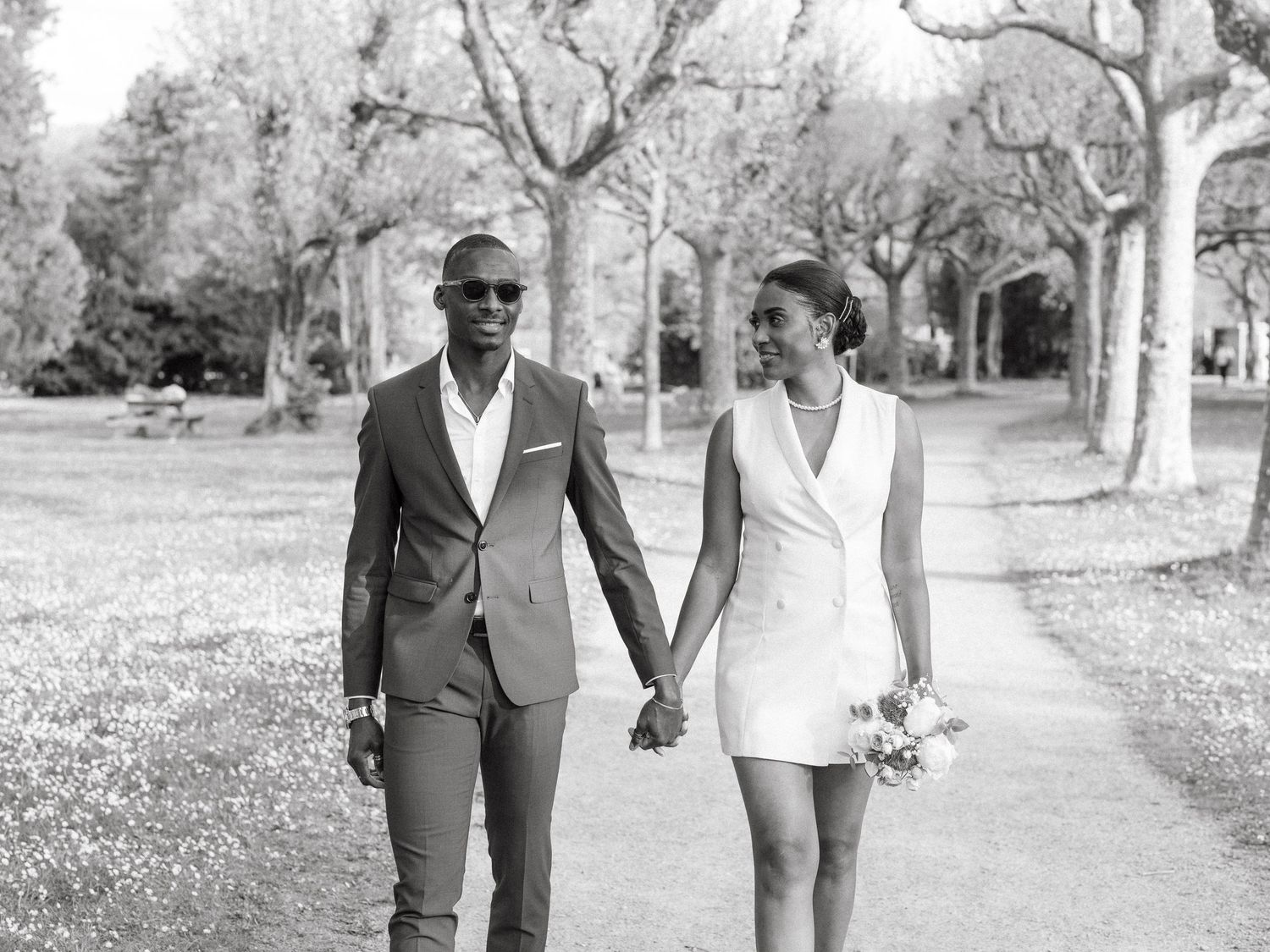 Black and white photo of smiling newlywed couple holding hands while strolling through a park.