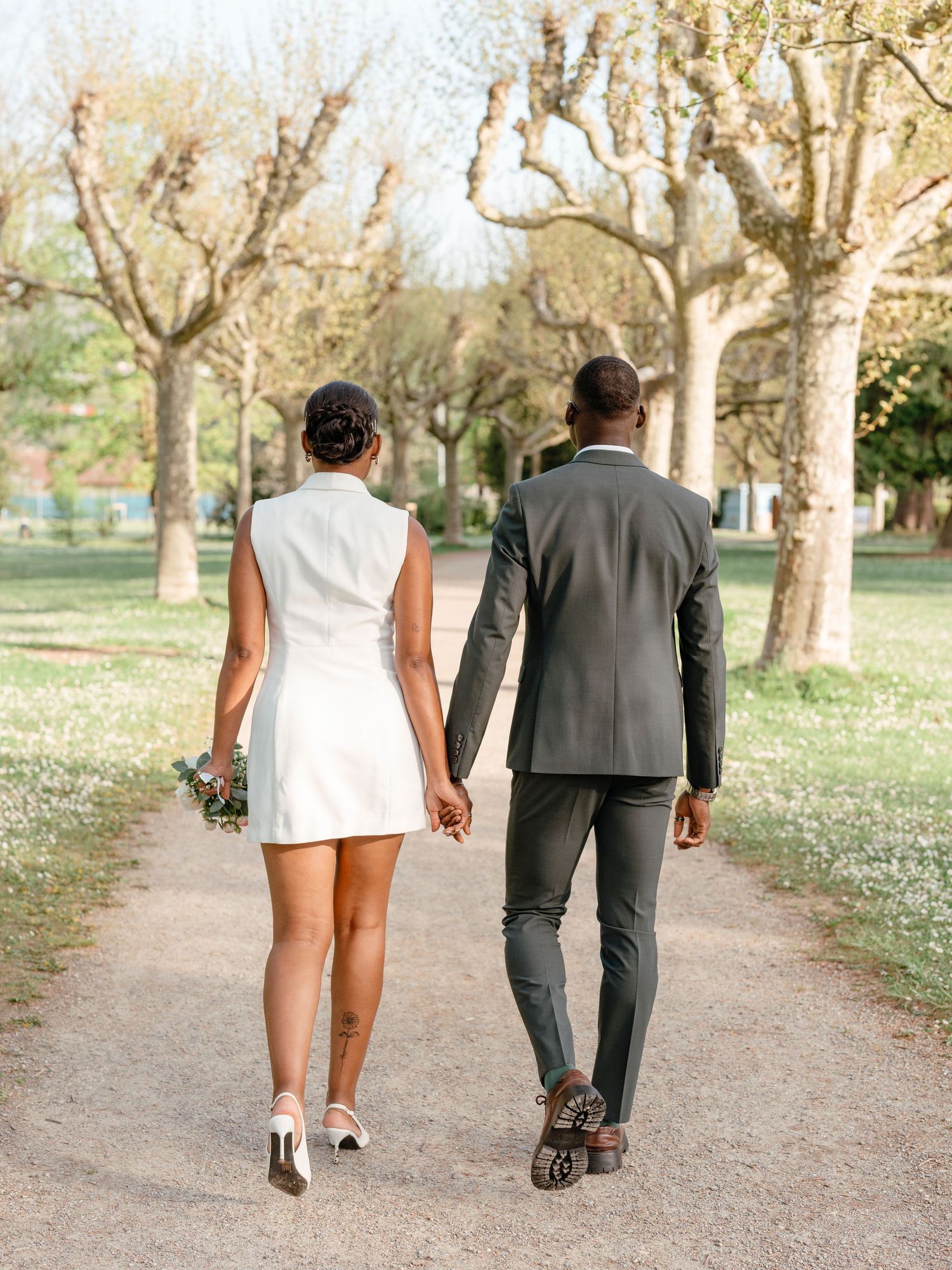 Newlywed couple walking away hand in hand down a scenic park path, bride in white mini dress.