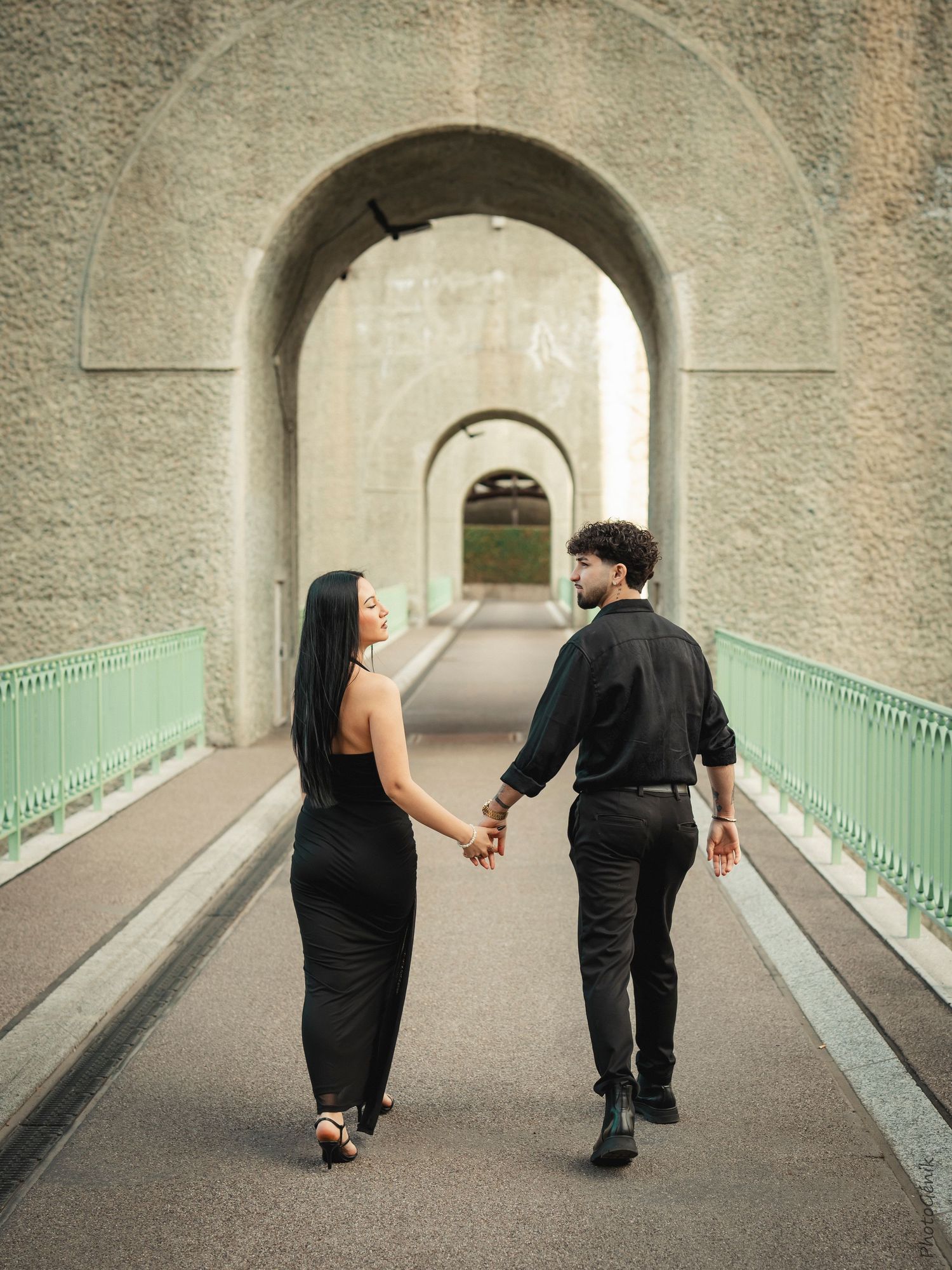 Couple in black outfits holding hands walking through arched bridge tunnel on a romantic stroll.