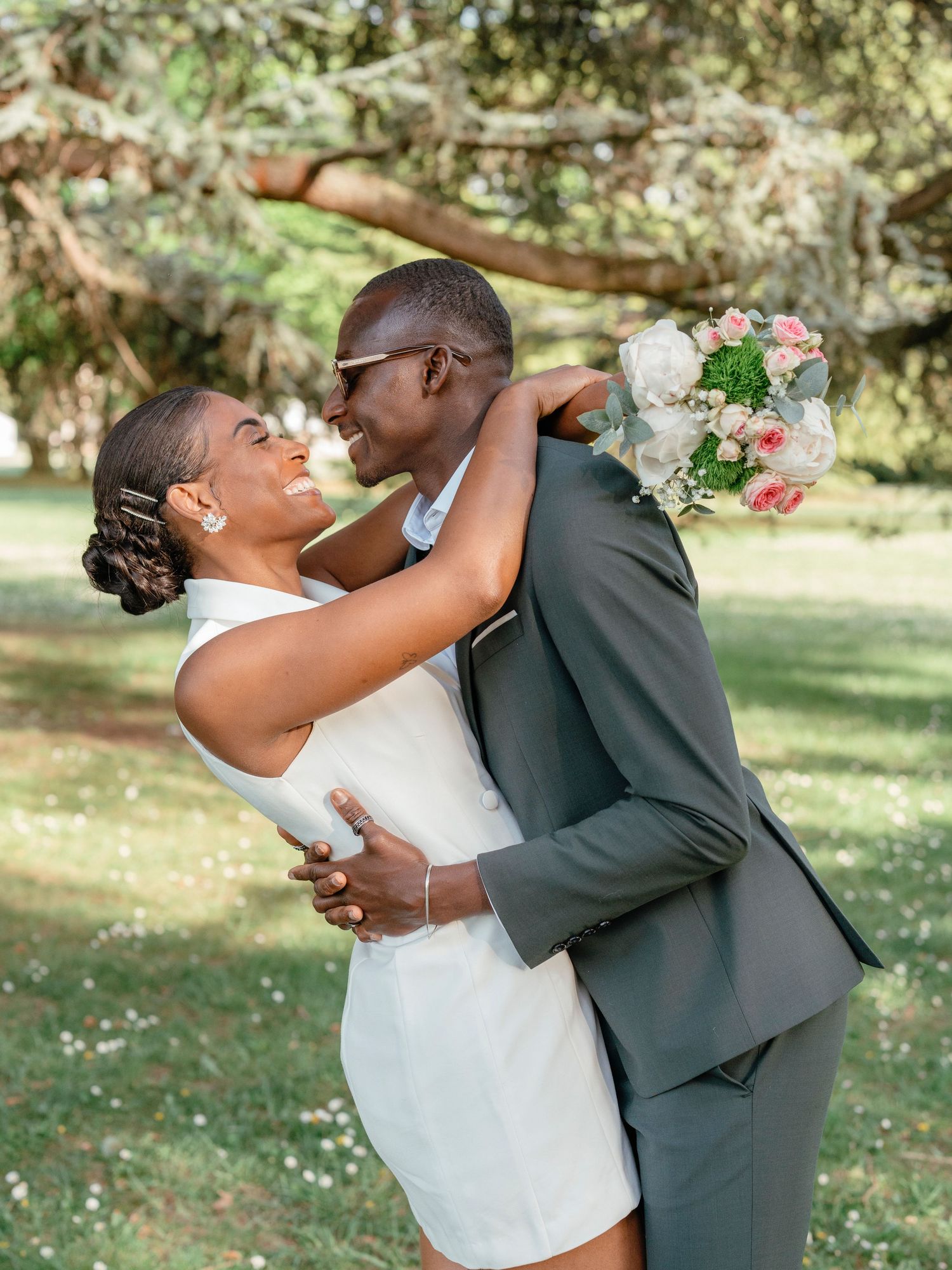 Joyful newlywed couple sharing a romantic moment, groom lifting bride in a white mini dress in a sunny park.