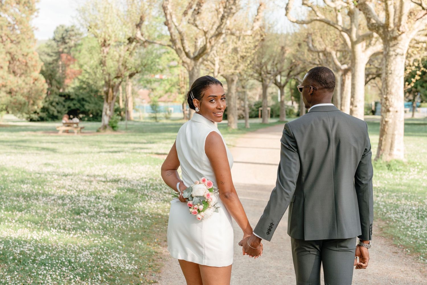 Bride glancing back with a smile while holding groom's hand and bouquet in a sunny park.