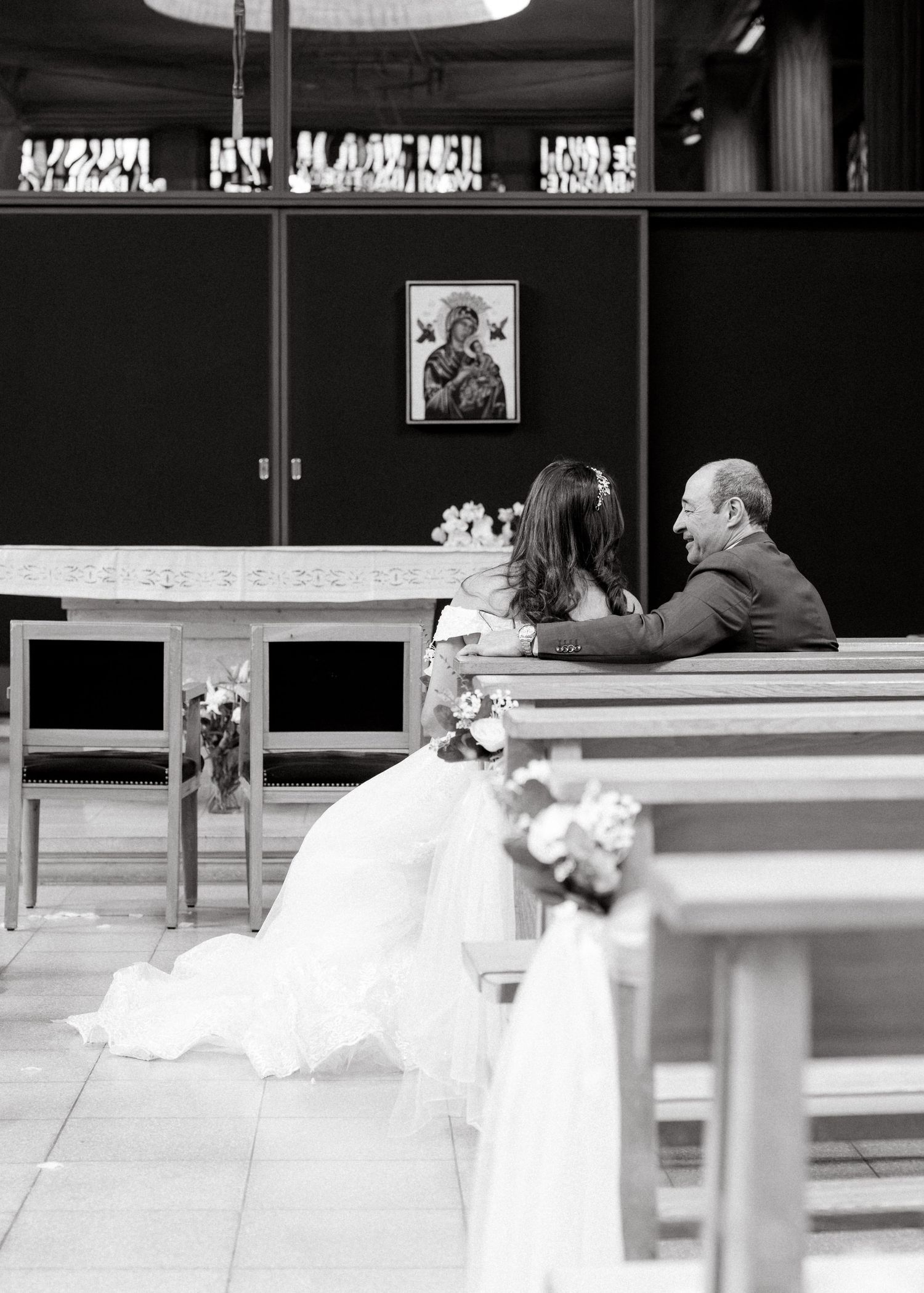 Black and white wedding photo of bride and groom sitting together in church pews during ceremony.