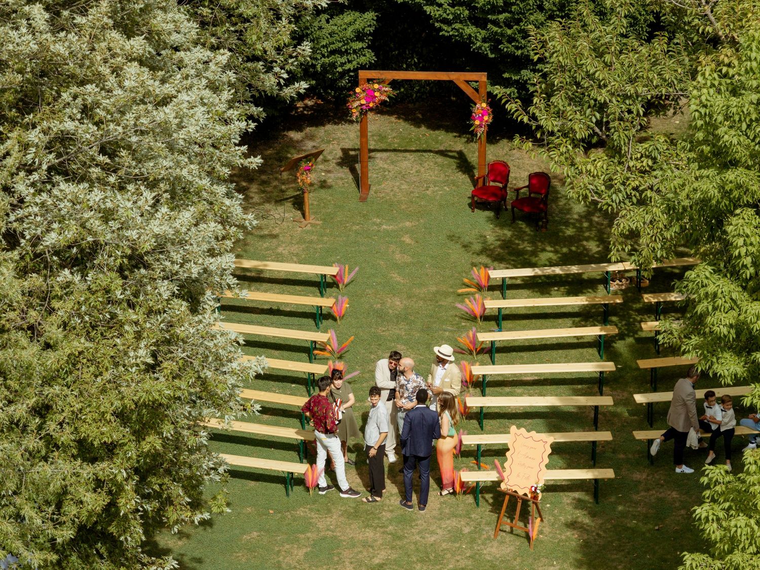 Aerial view of an outdoor wedding ceremony setup with wooden benches, floral arch, and guests on a green lawn.
