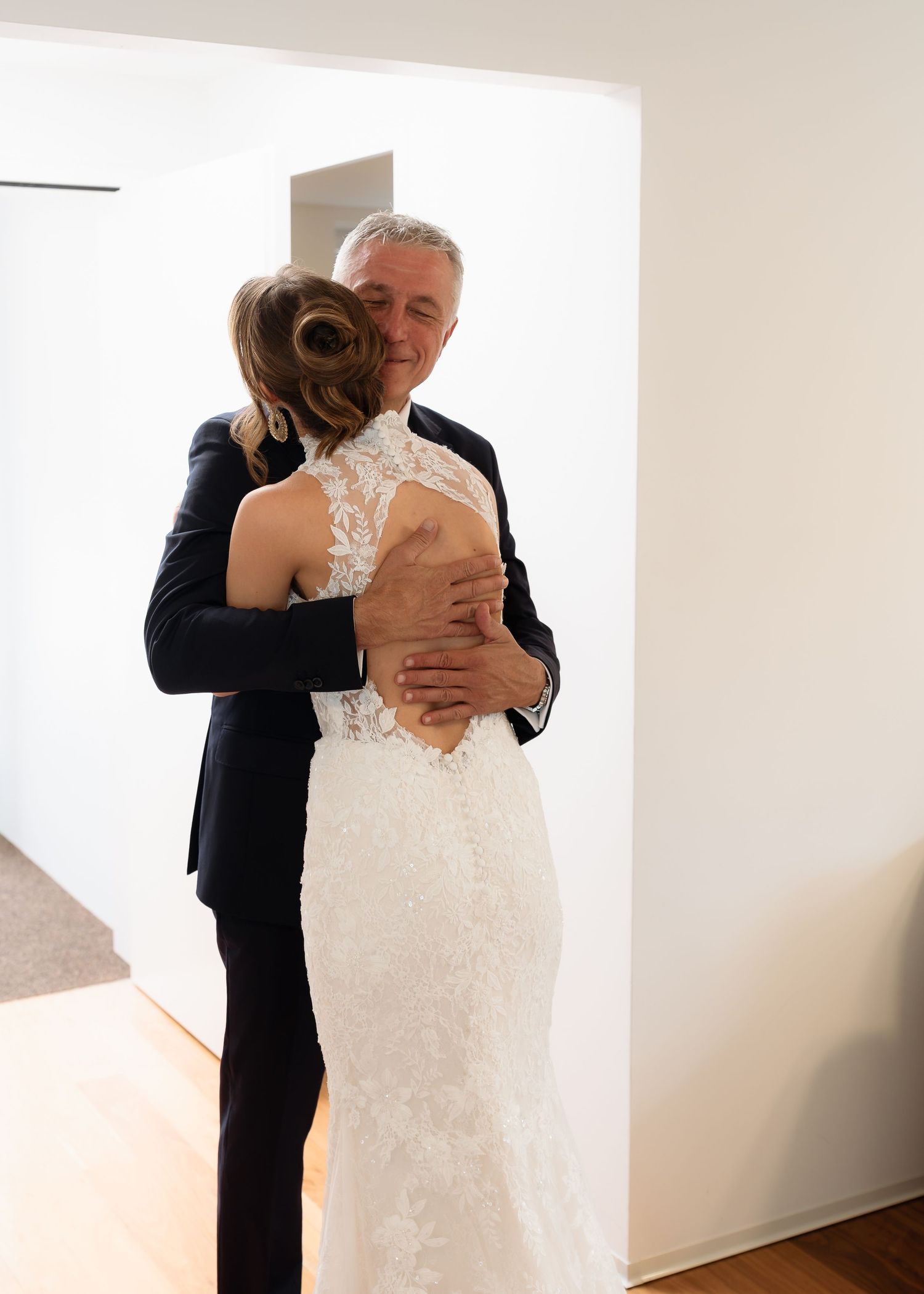 Bride in lace wedding gown embracing a man in a dark suit during an emotional pre-wedding moment indoors.