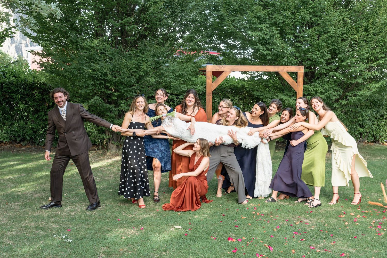 Wedding party playfully lifts bride outdoors near wooden arch surrounded by smiling guests in garden setting.