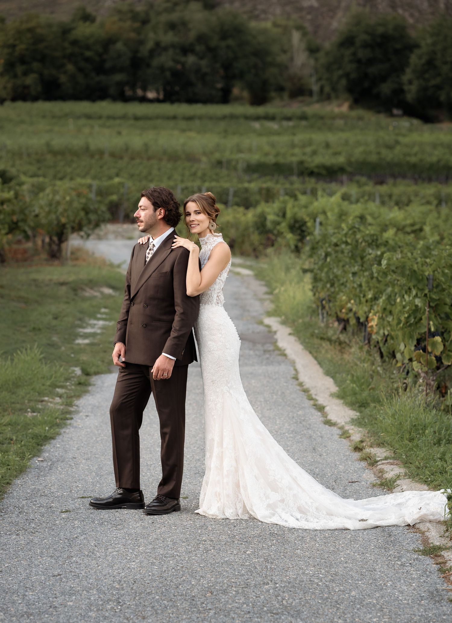 Bride and groom pose on a vineyard path, she in a lace gown with train, he in a brown suit.
