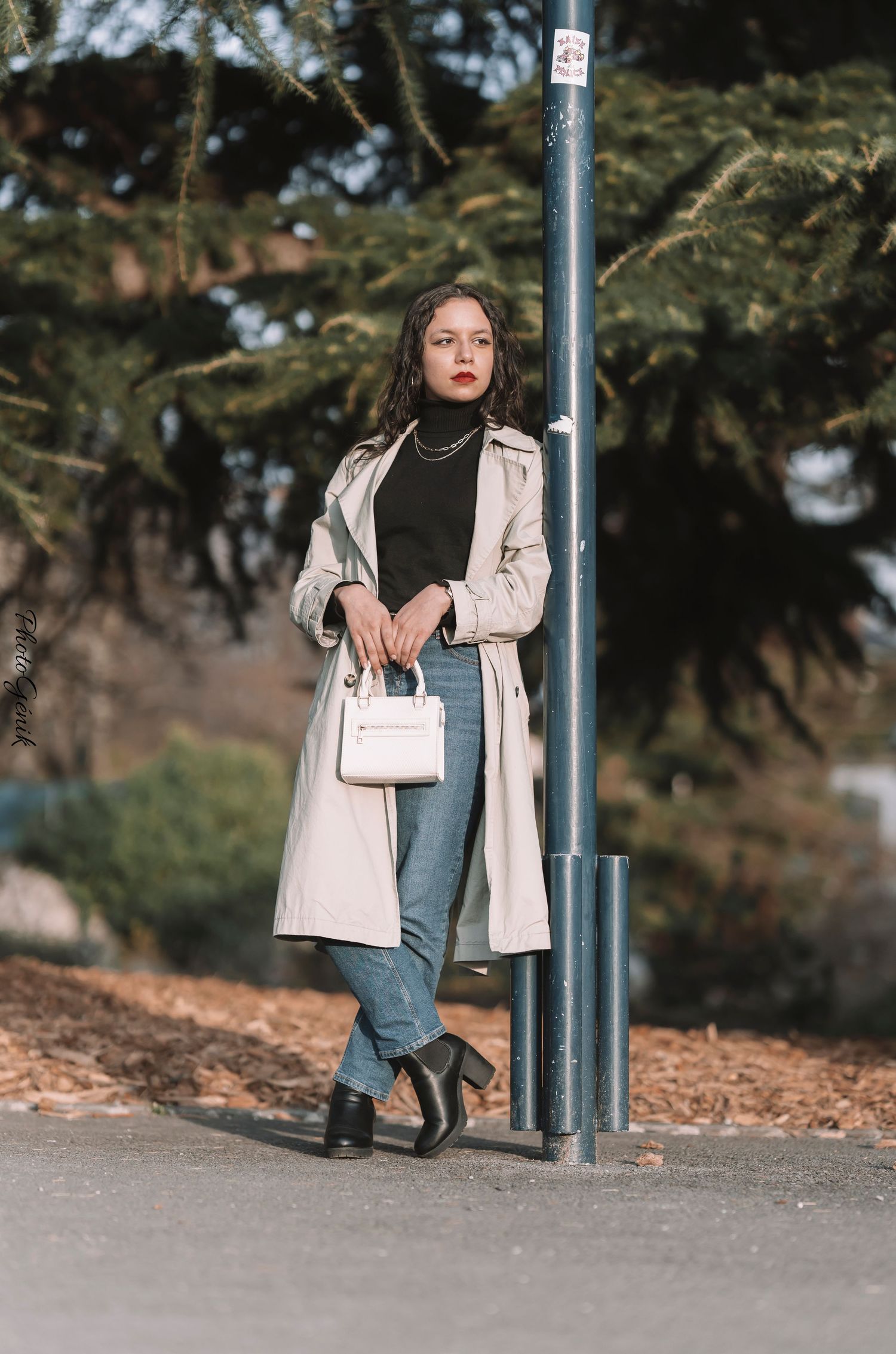 Young woman in beige trench coat, black turtleneck, jeans, and ankle boots, holding white bag while leaning on pole outdoors.