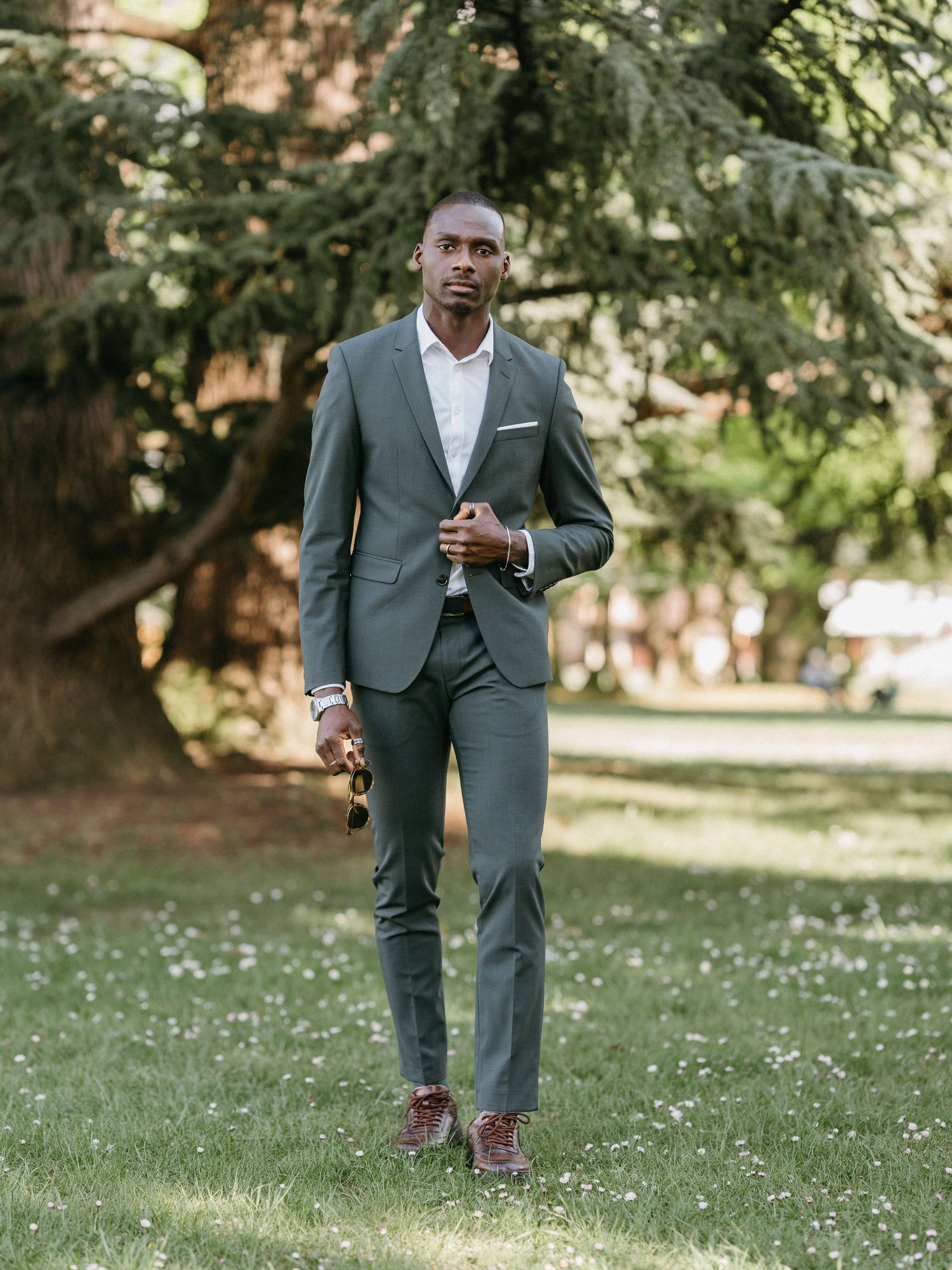 Stylish groom in tailored grey suit and brown shoes strolling through sunlit park with tall trees.