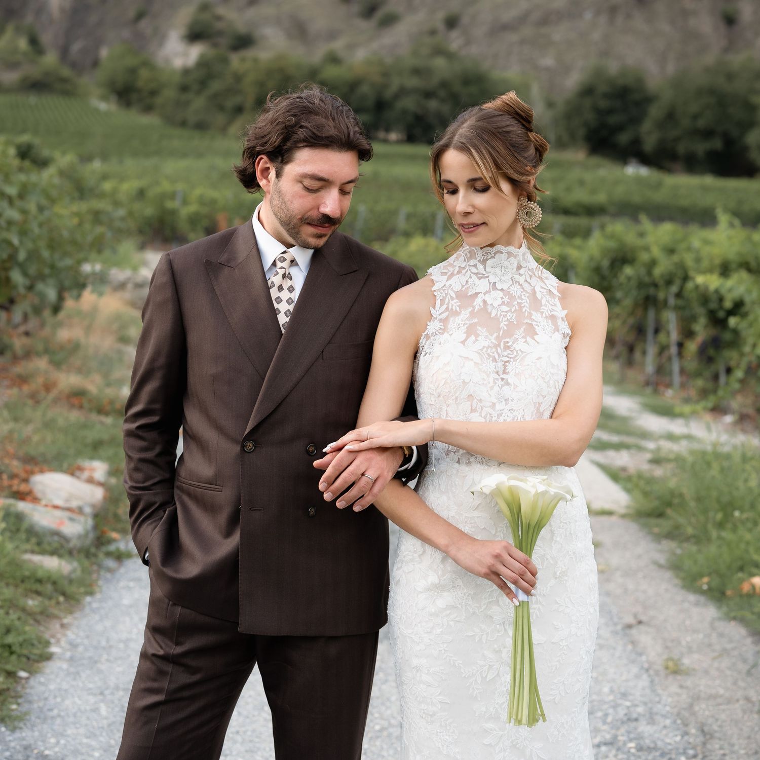Bride in lace halter gown holds calla lilies beside groom in brown suit, posing on vineyard path.