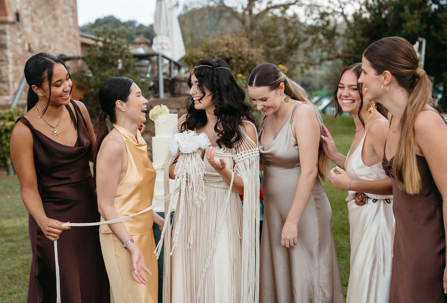 Bride in white gown laughing with six bridesmaids in brown and gold dresses outdoors at a wedding celebration.