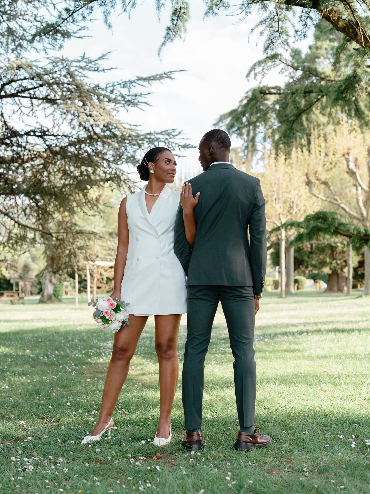A bride in a white blazer mini dress and groom in a dark suit pose back-to-back in a scenic park.
