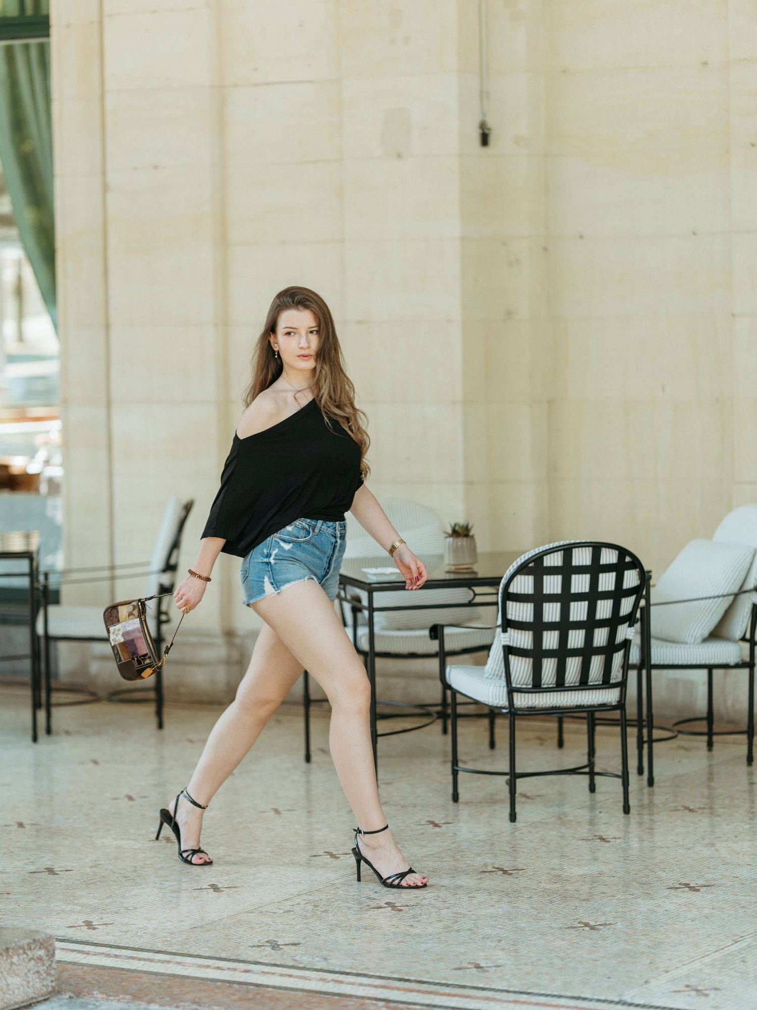 Young woman in black top and denim shorts walking in strappy heels at outdoor venue.