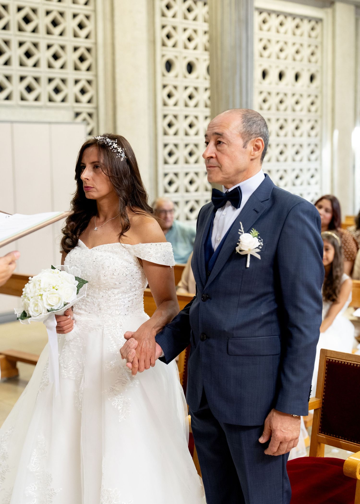Bride in white off-shoulder gown holding bouquet walks down church aisle with man in navy suit at wedding ceremony.