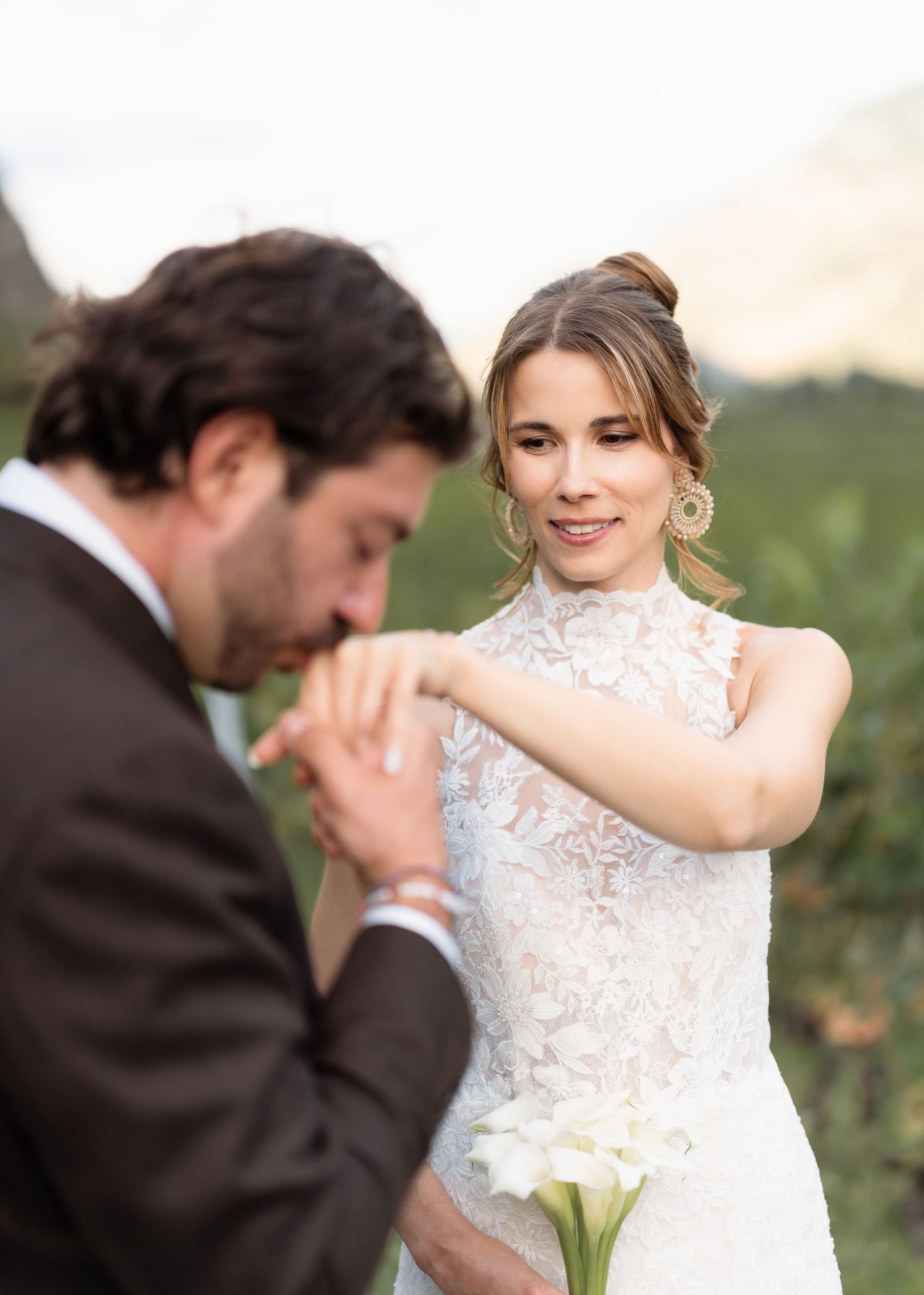 A bride in a lace wedding dress holds white calla lilies as the groom kisses her hand during an outdoor ceremony.