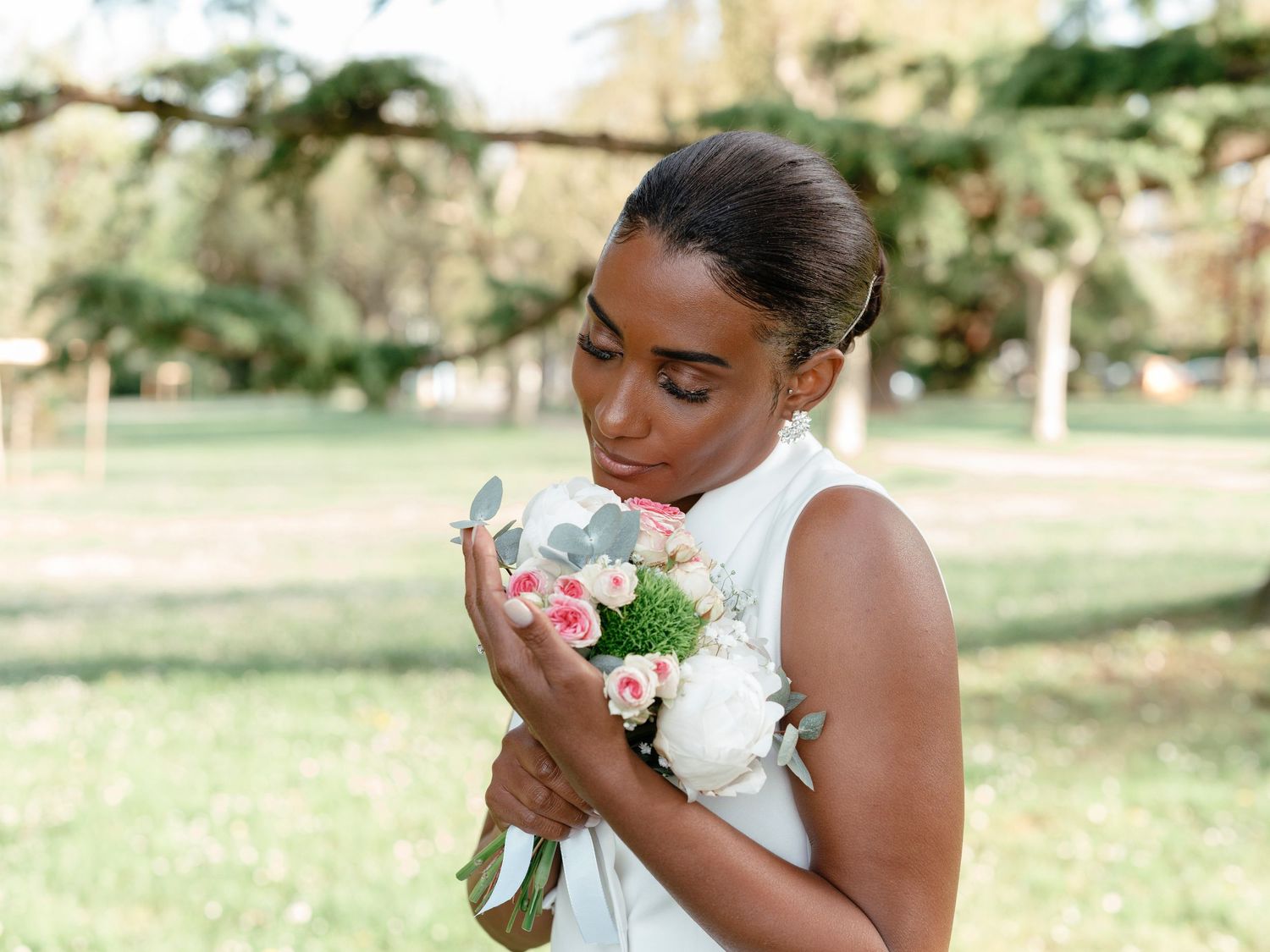 Beautiful Black bride smelling her pink and white wedding bouquet in a sunny park setting.