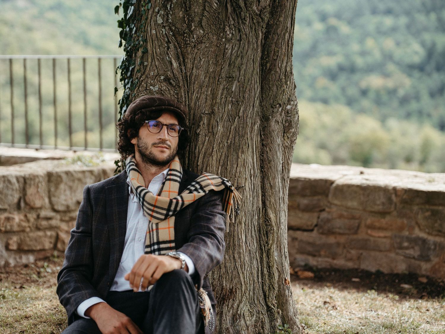 Stylish man in a plaid blazer and Burberry scarf sitting against a tree in a scenic countryside setting.