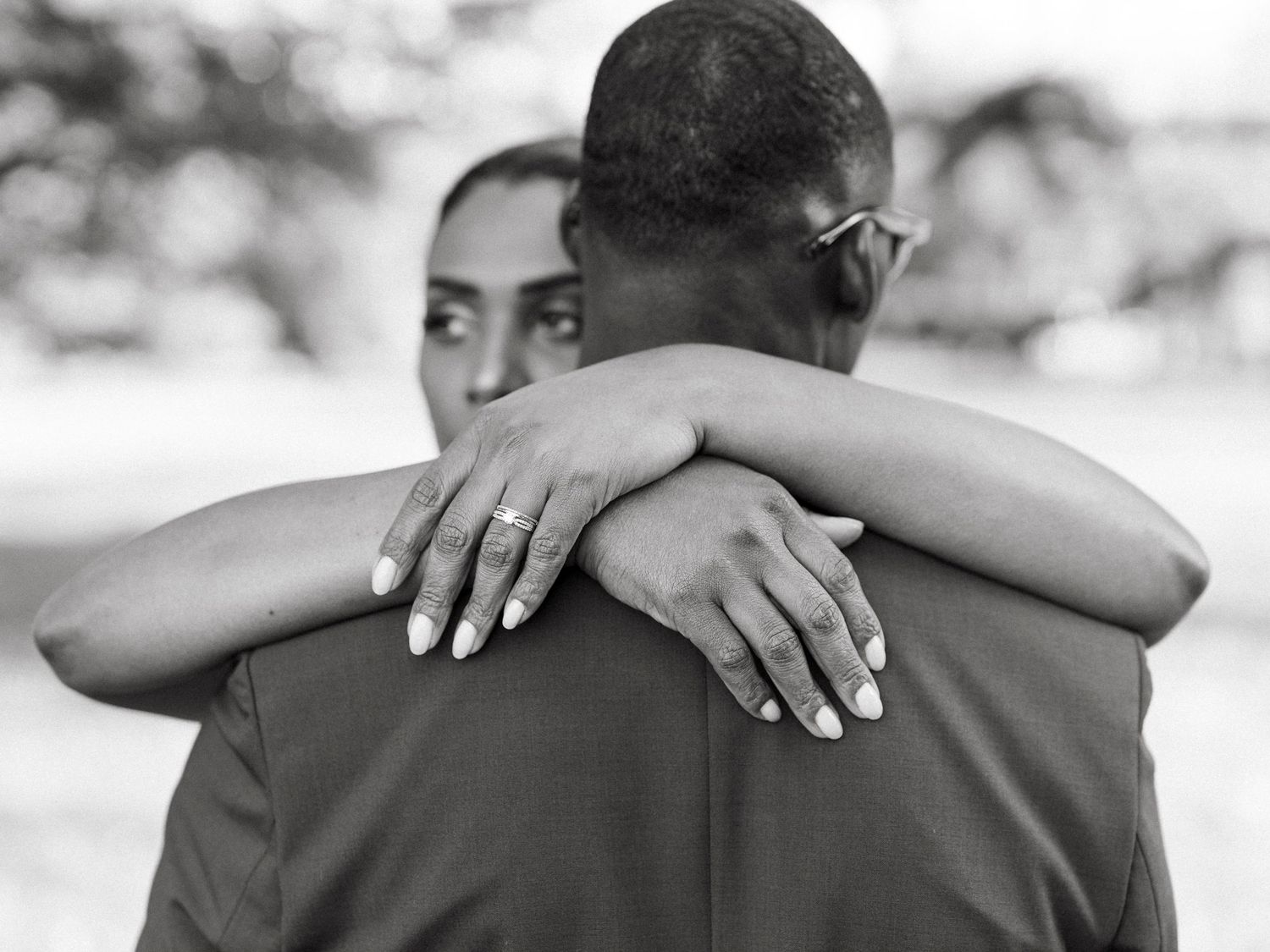Black and white close-up of bride hugging groom, showcasing her diamond engagement and wedding rings on his back.