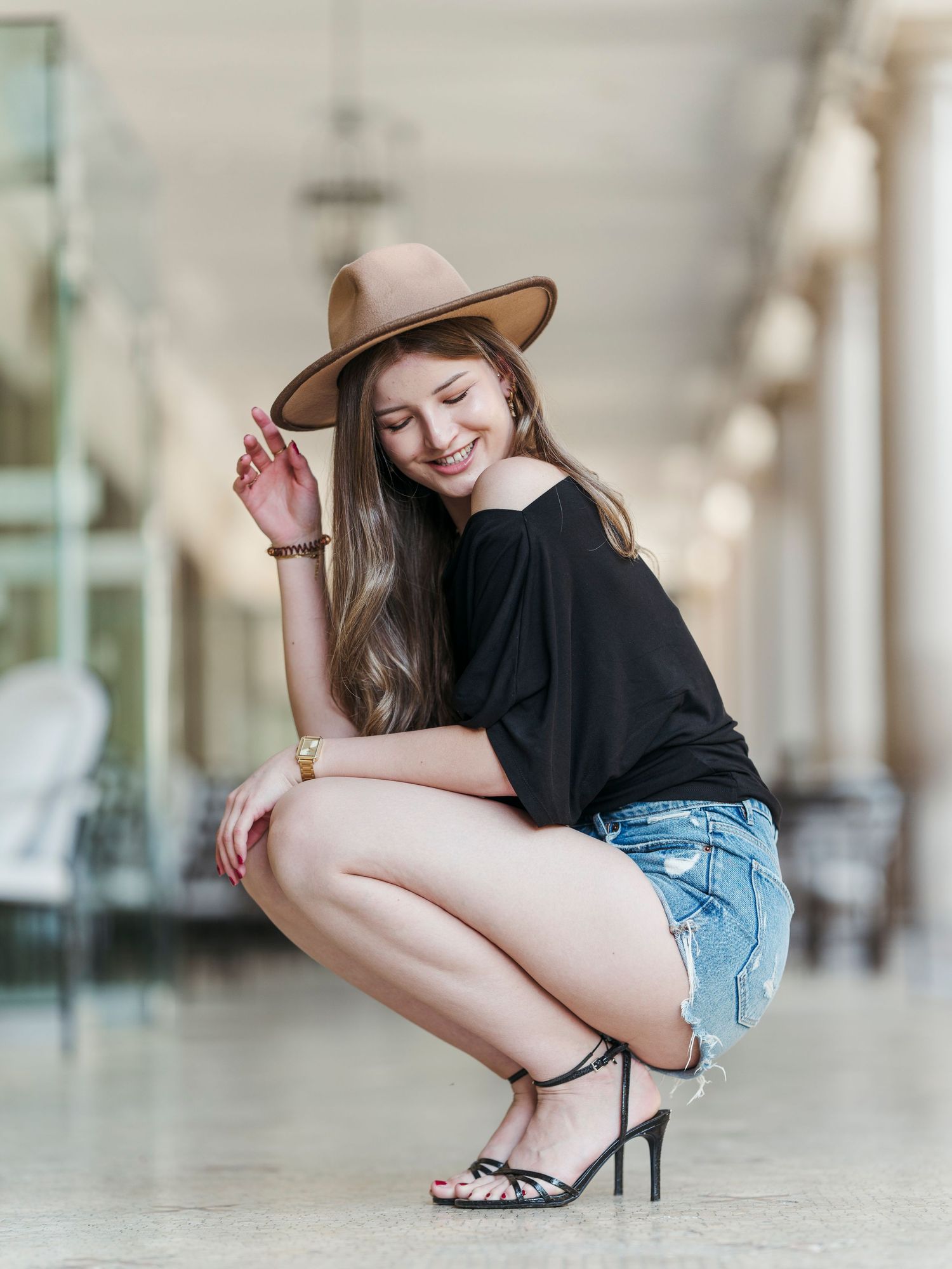 Smiling woman in denim shorts, black top, and tan wide-brim hat squatting gracefully in a blurred corridor.