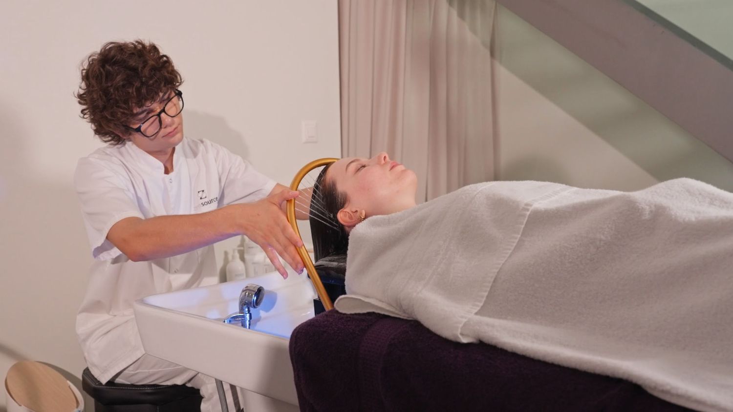 Professional hairdresser washing client hair at salon shampoo bowl, client reclined with white towel draped over shoulders.