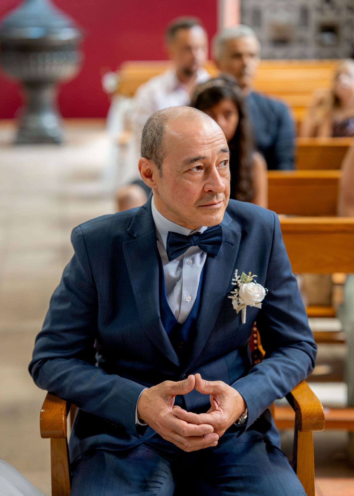 Elegant man in navy blue suit with bow tie and boutonniere sits in church pew at a wedding ceremony.