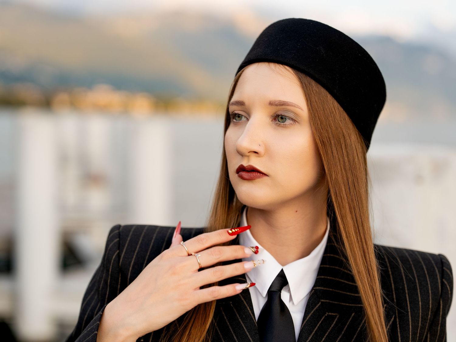 Young woman in pinstripe blazer and black beret with dark red lips and manicured nails posing outdoors.