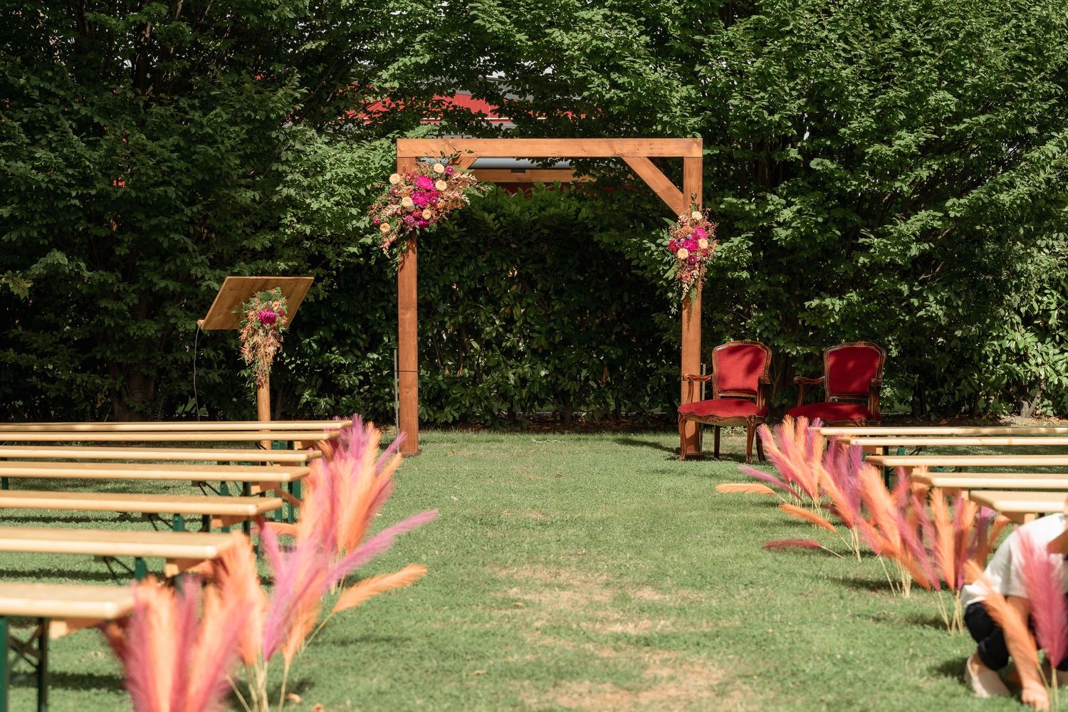 Outdoor wedding ceremony setup with wooden arch adorned with pink flowers, lined benches, and pink grass along the aisle.