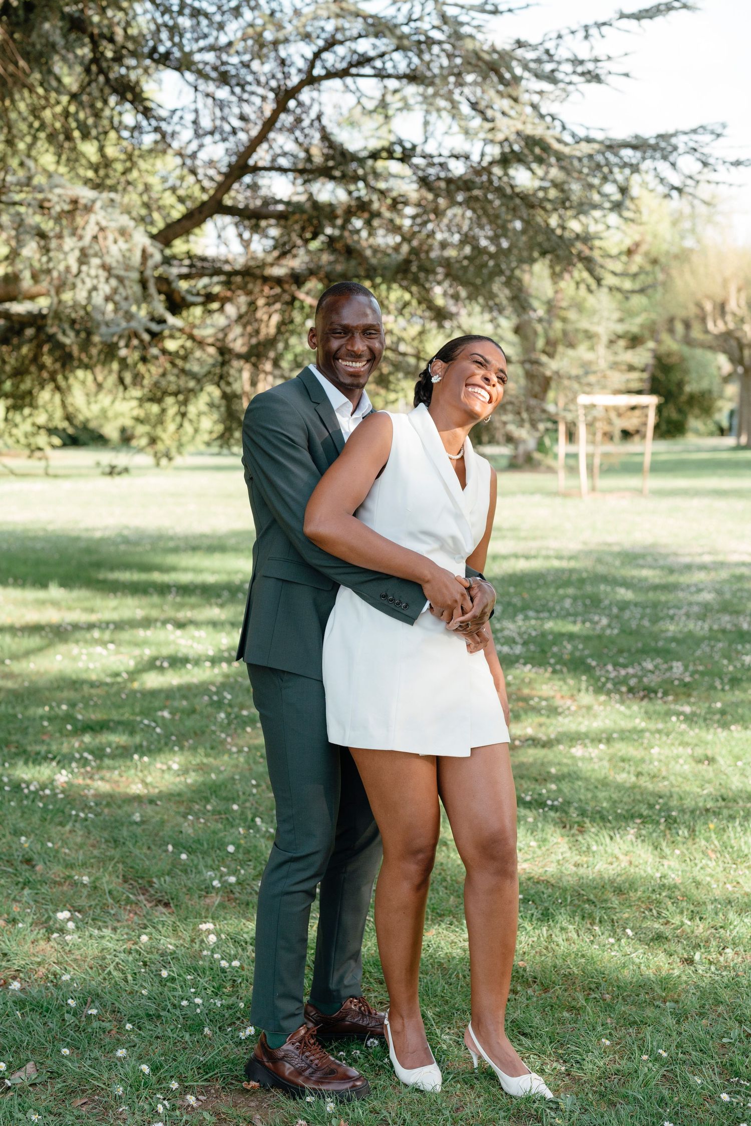 A joyful couple in wedding attire embrace and laugh together in a lush green park on a sunny day.