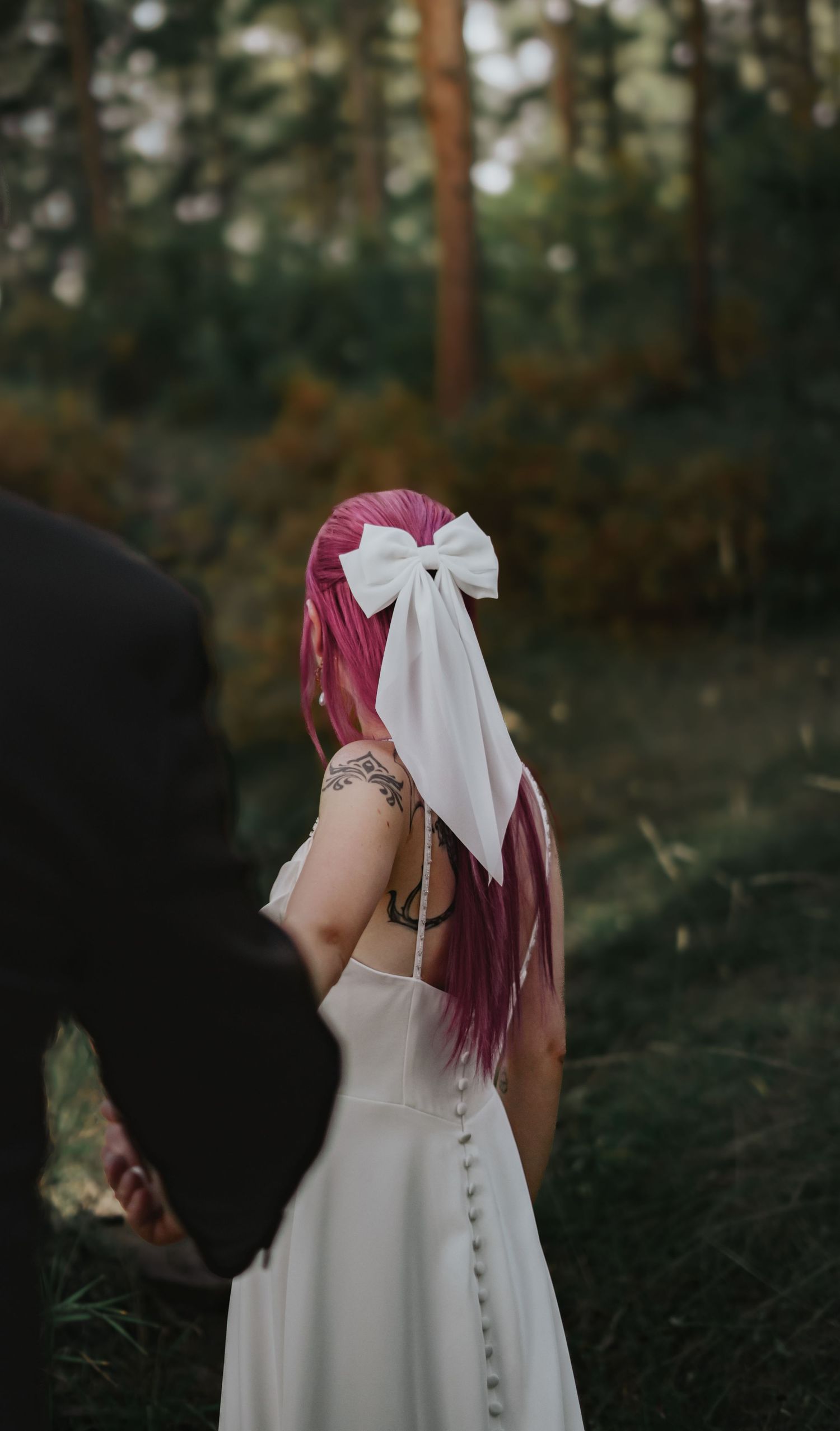 A bride with pink hair stands in a forest wearing a classic white wedding dress while reaching out to her husband in Pueblo Mountain Park.