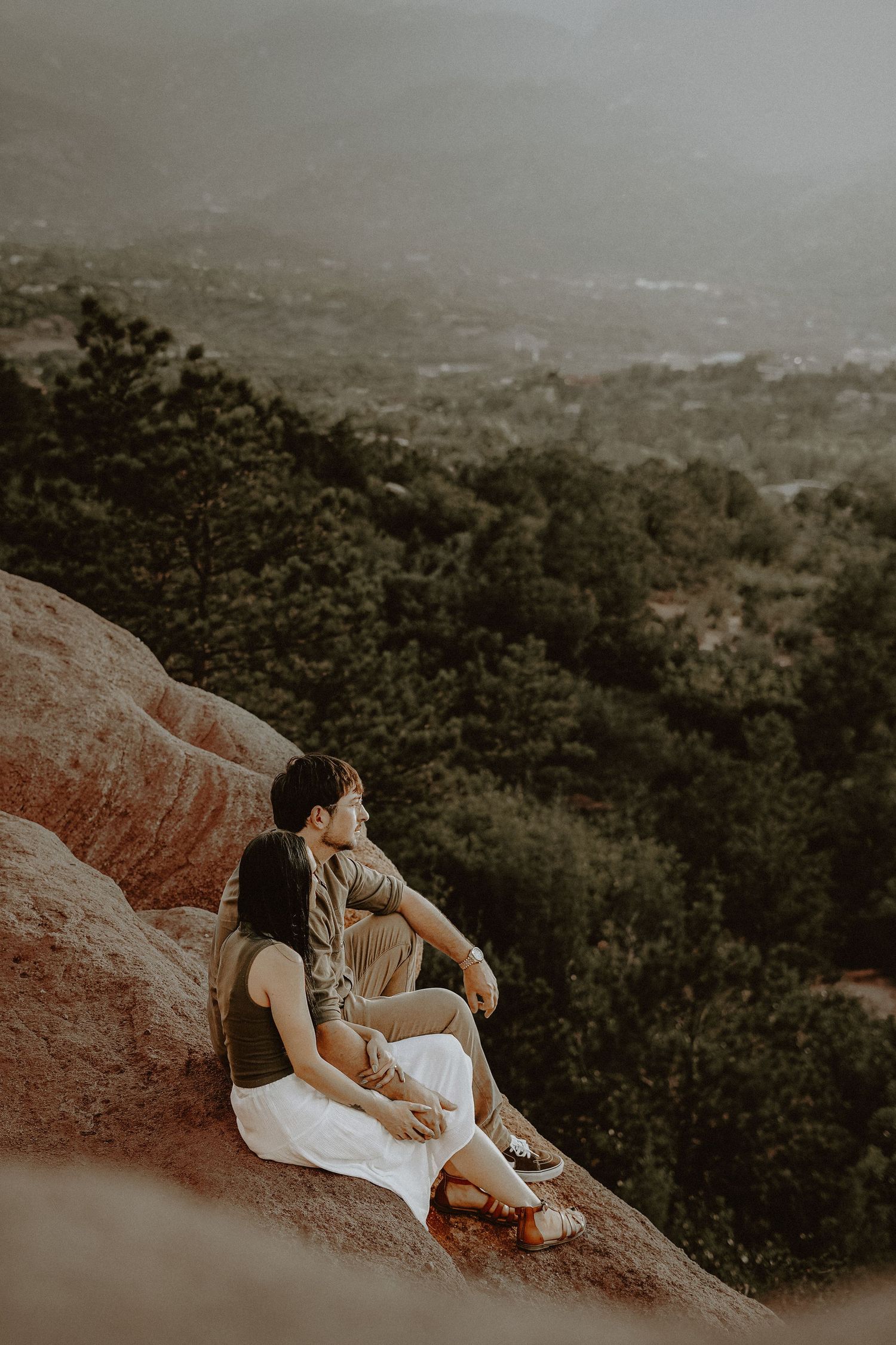 Couple in neutral attire relaxing on red rocks overlooking vast mountain valley vista at Garden of the Gods.