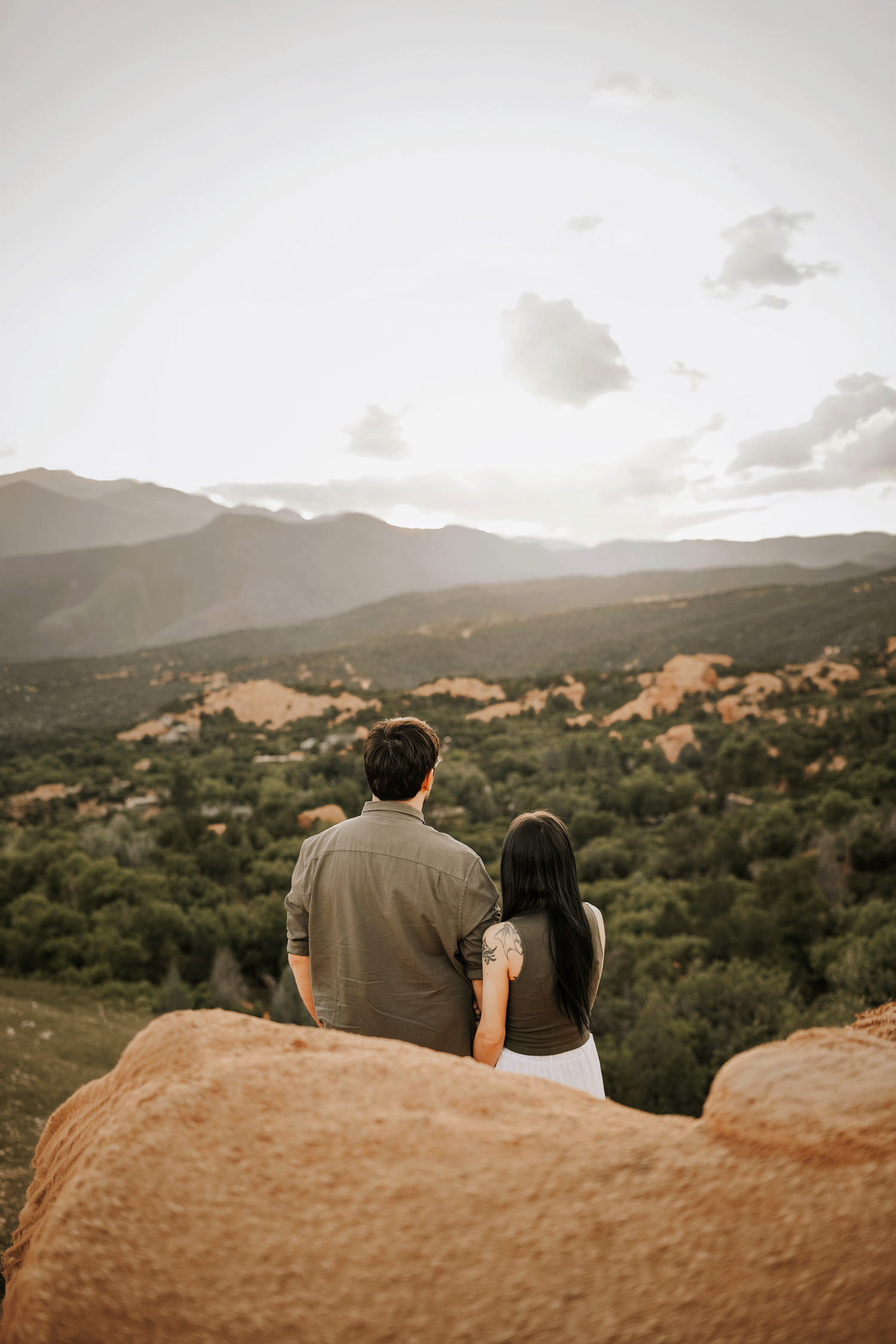 Newly engaged couple sits together on red rocks overlooking Garden of the Gods mountain valley at sunset.