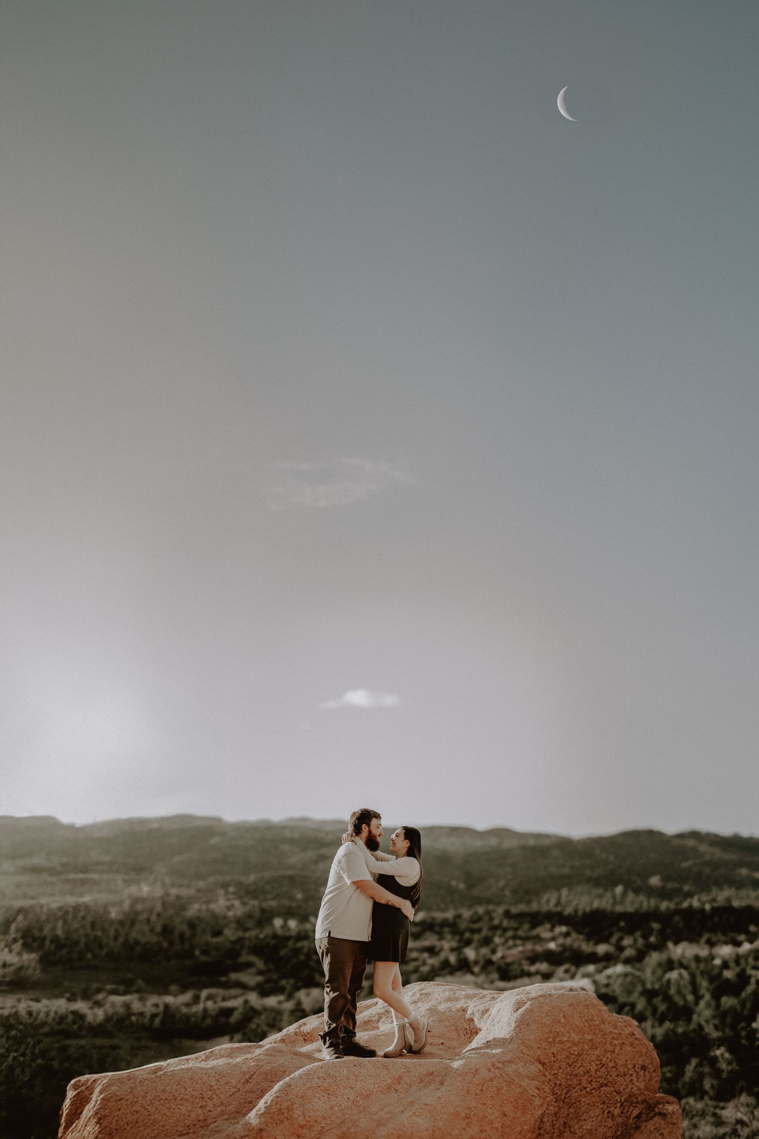 Couple embraces on Garden of the Gods red rocks under crescent moon in mountain landscape.