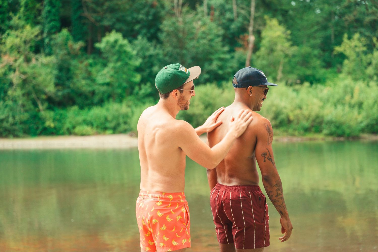Two shirtless men in swim trunks and caps stand by a lake, one placing hand on the other's shoulder.