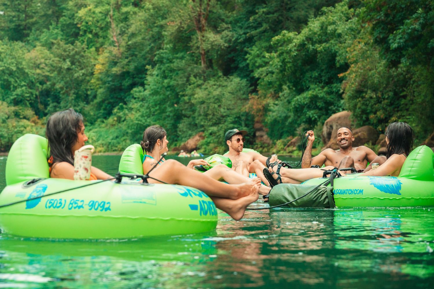 Group of friends relaxing on green inflatable tubes floating on a calm river surrounded by lush green trees.