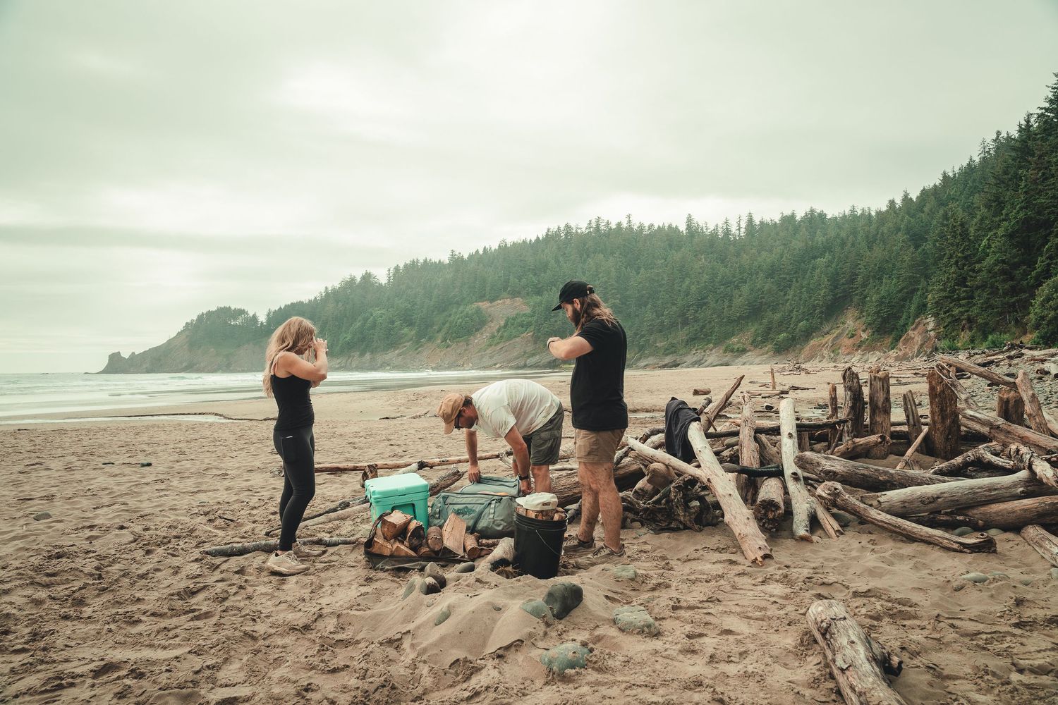 Three people gather driftwood on a rugged Pacific Northwest beach surrounded by logs with forested hills in the background.