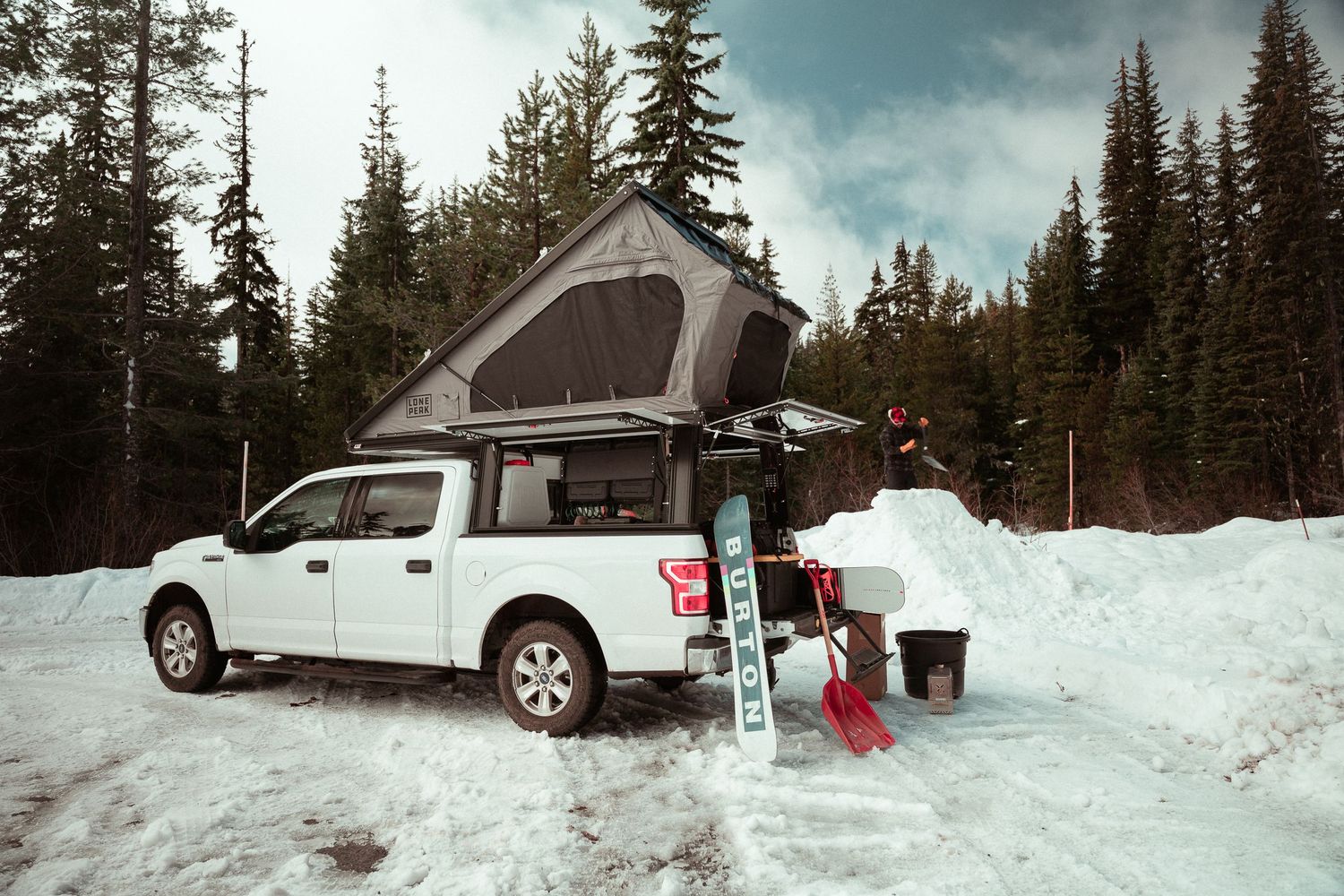 White pickup truck with rooftop tent camping in snowy winter forest with ski equipment and gear setup.