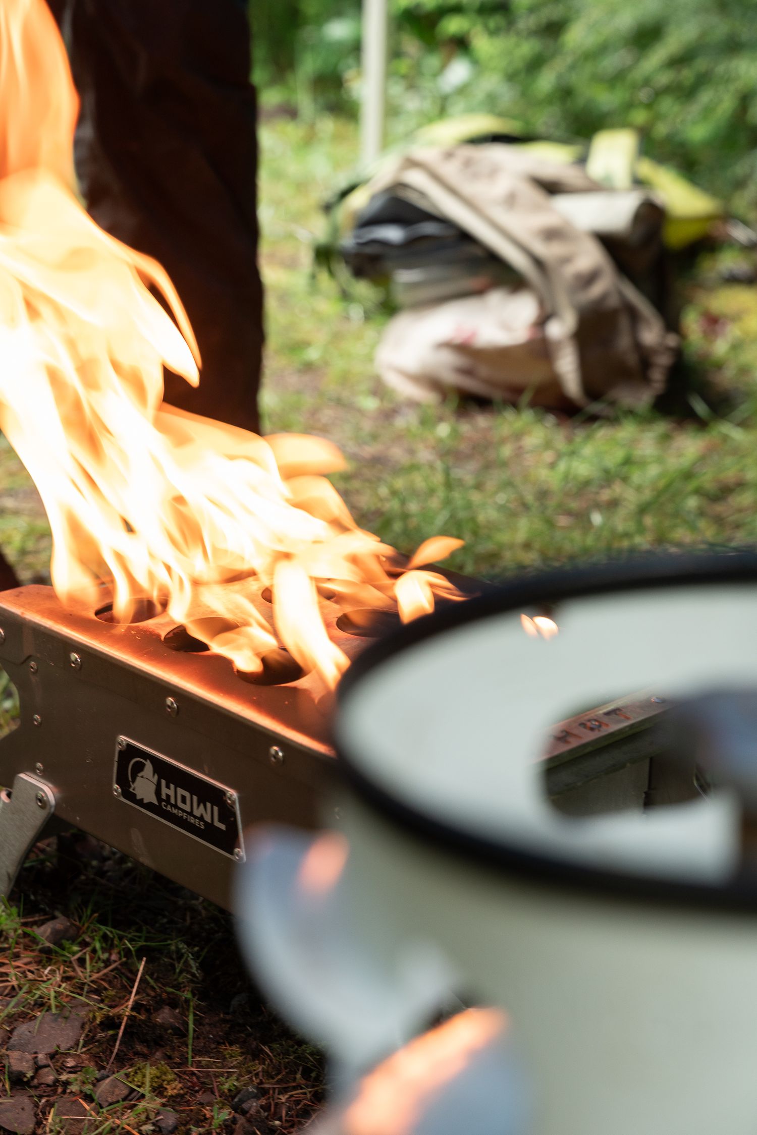 HOWL camp stove with large flames burning outdoors, enamel mug in foreground, backpacks visible in background.