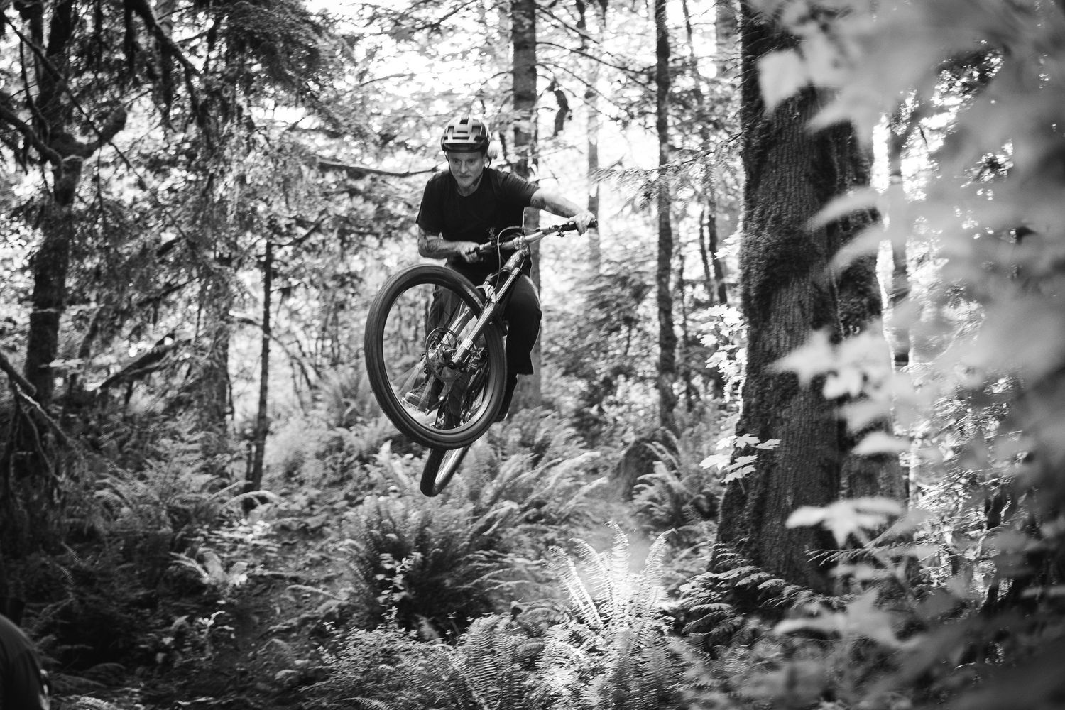 Mountain biker performing a jump trick through a lush forest trail in black and white photography.